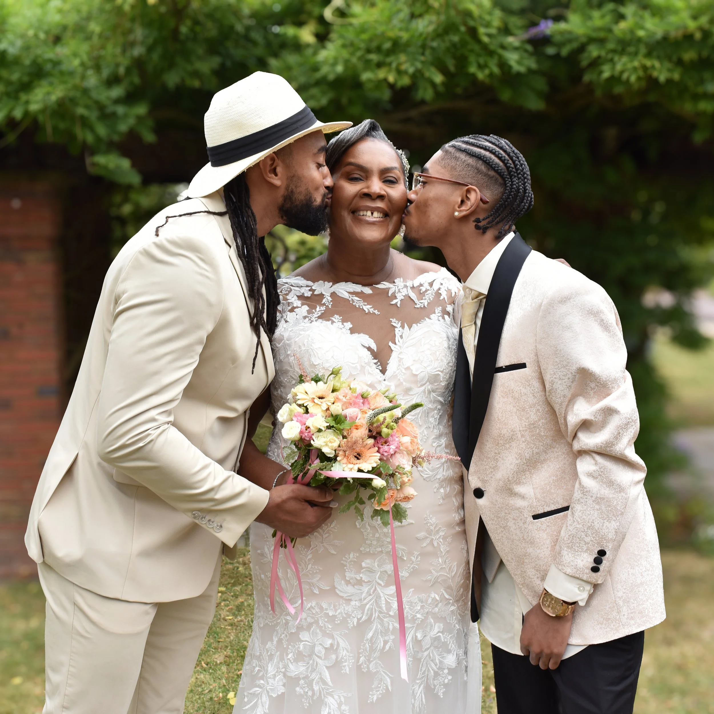 A wedding scene with a bride holding a bouquet of flowers, being kissed on the cheeks by two men dressed in light-colored suits, outdoors with green trees in the background.