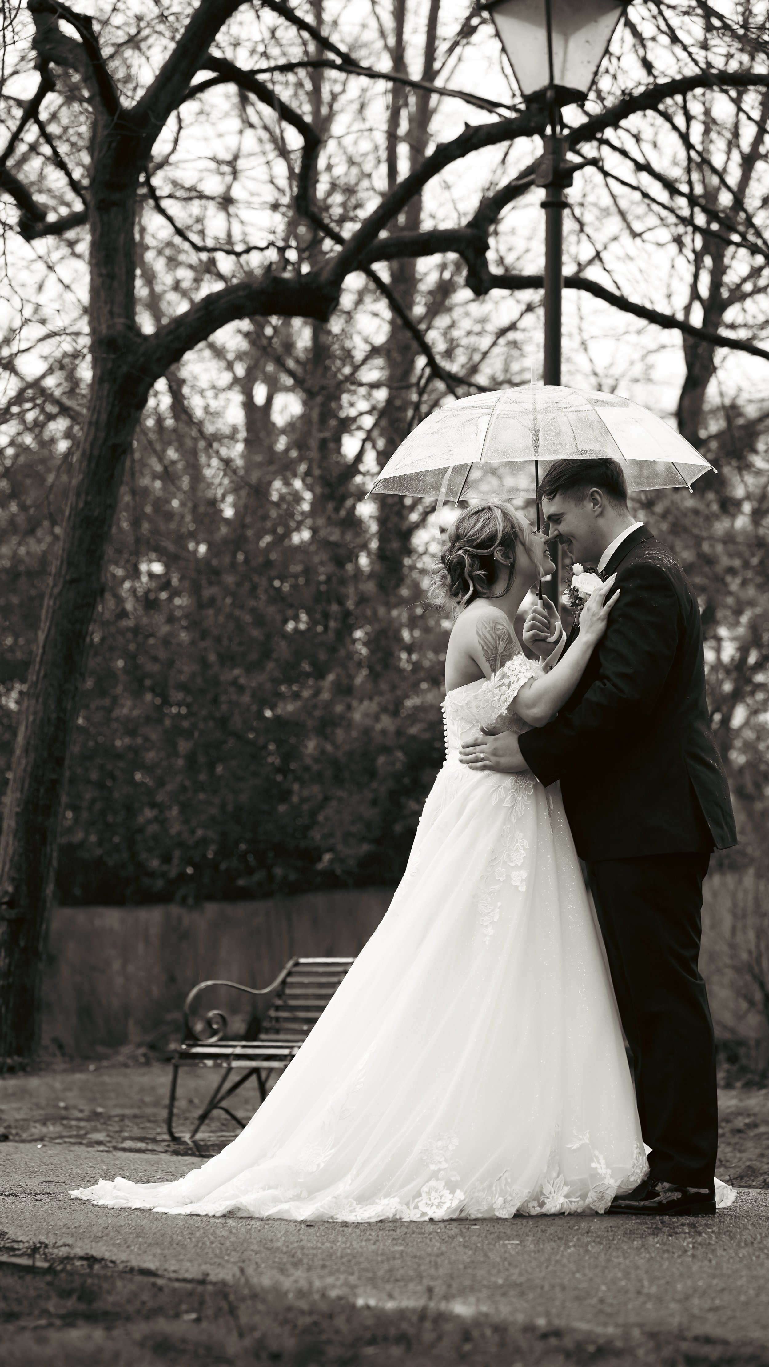 A wedding couple standing under an umbrella outdoors, gazing into each other's eyes. The bride is in a white wedding dress, and the groom is in a black suit. The background features trees and a park bench.
