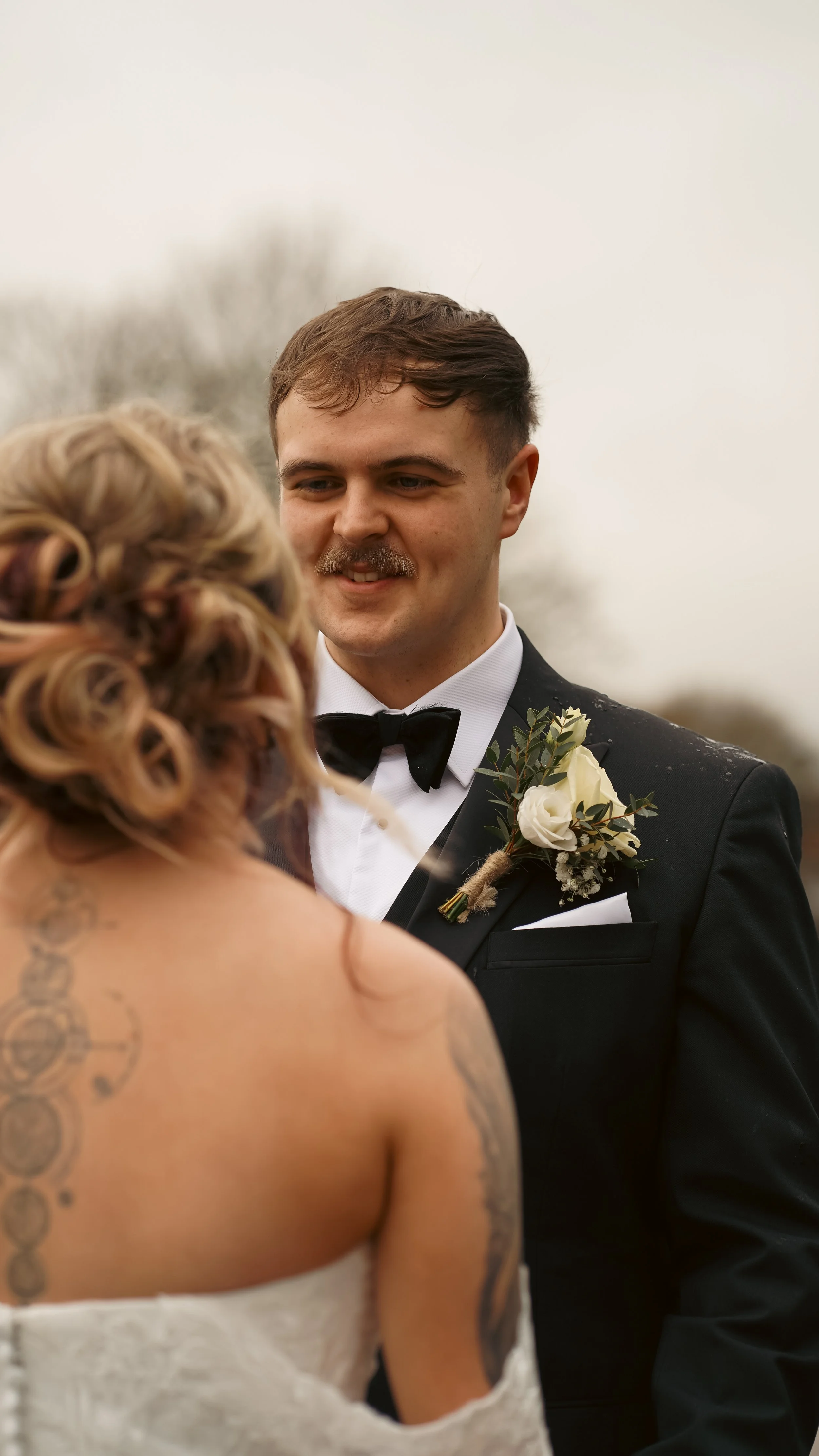A groom in a black tuxedo and bow tie stands outdoors, smiling at a bride with curly hair and tattoos, during a wedding ceremony.