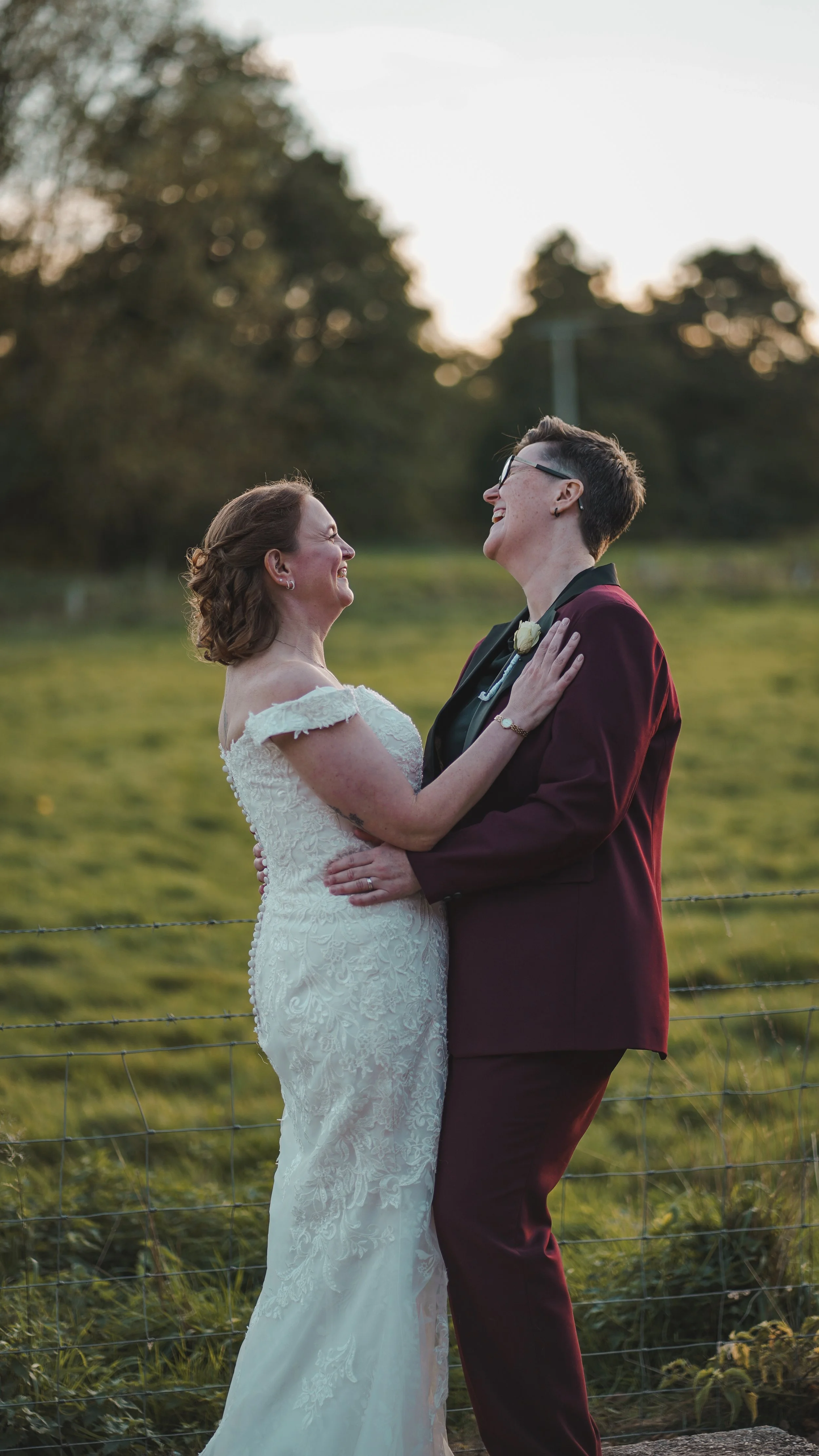 Two women, one in a wedding dress and the other in a burgundy suit, share a joyful moment outdoors during sunset, holding each other and smiling.