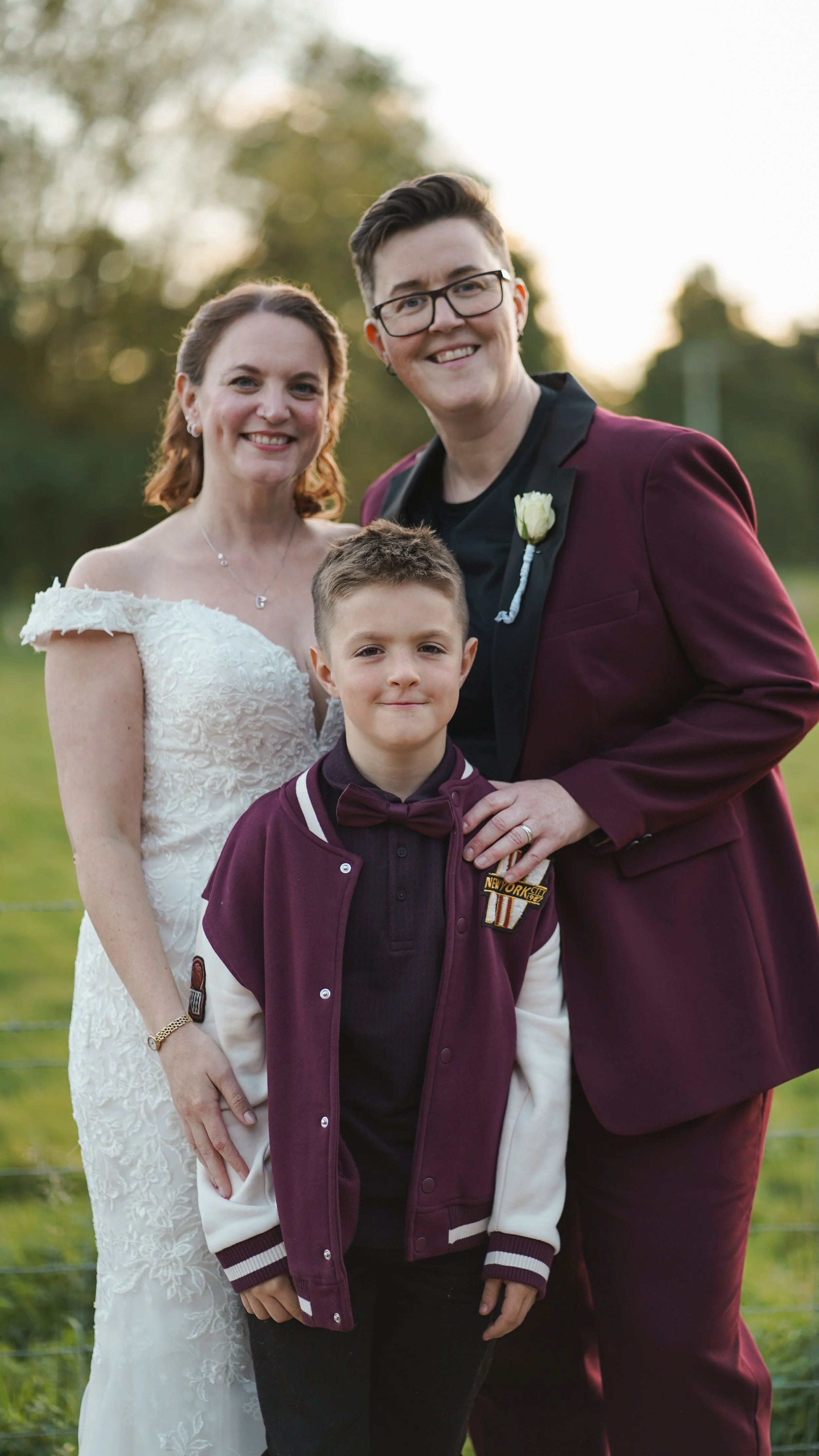A young woman in a white wedding gown and a woman with short dark hair and glasses in a burgundy suit stand outdoors with a young boy in front, dressed in a sporty jacket, at sunset.