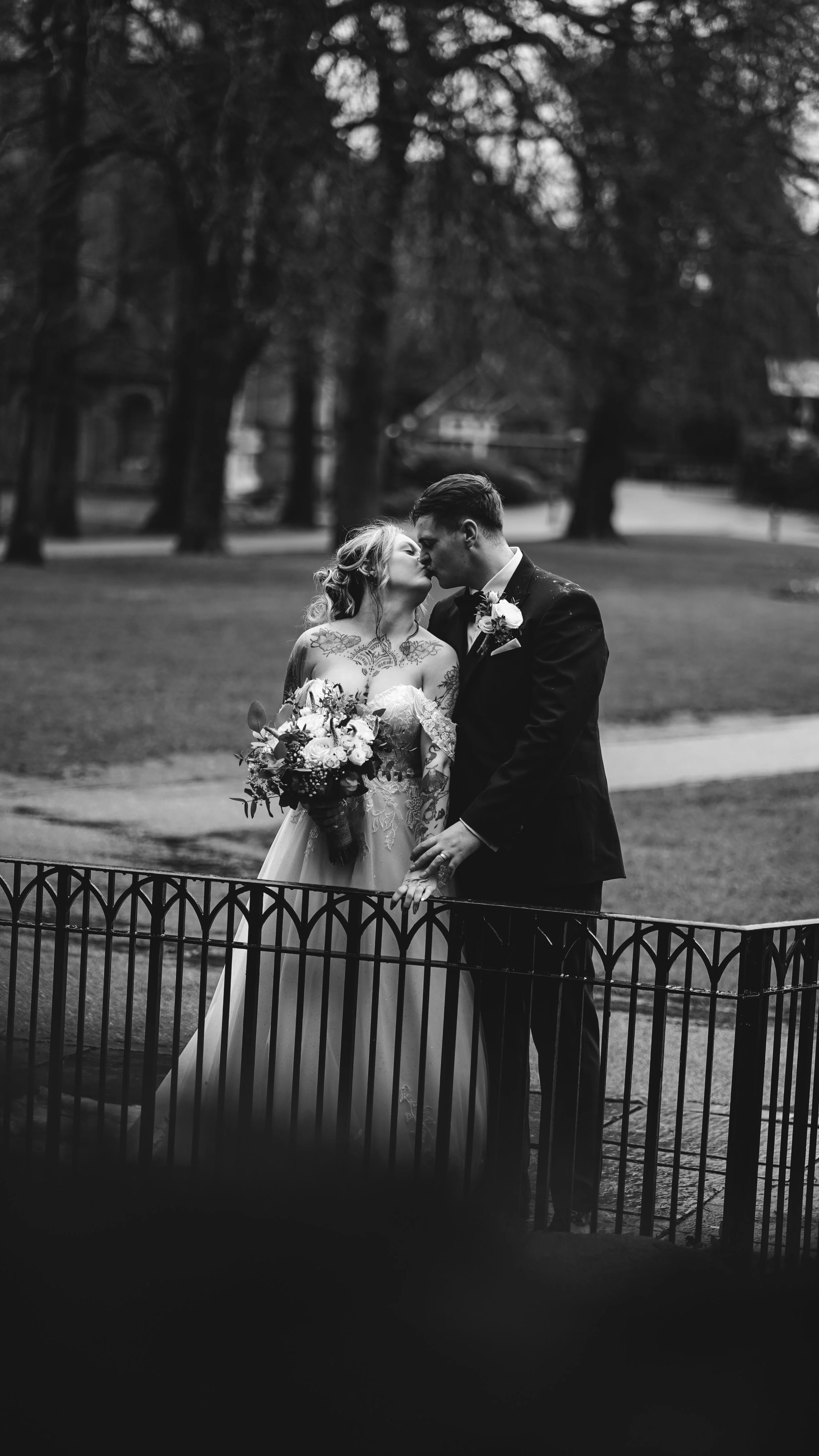 Black and white photo of a bride and groom sharing a kiss in a park, with the bride holding a bouquet of flowers and the groom wearing a tuxedo.