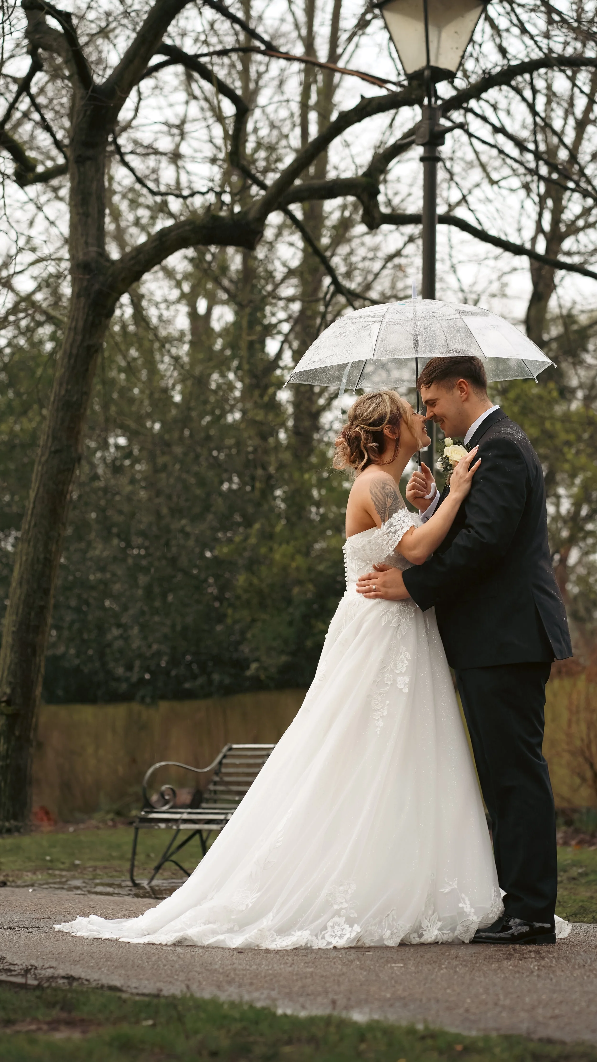 A bride and groom sharing a kiss under a clear umbrella outdoors on a rainy day, surrounded by trees and a park bench.