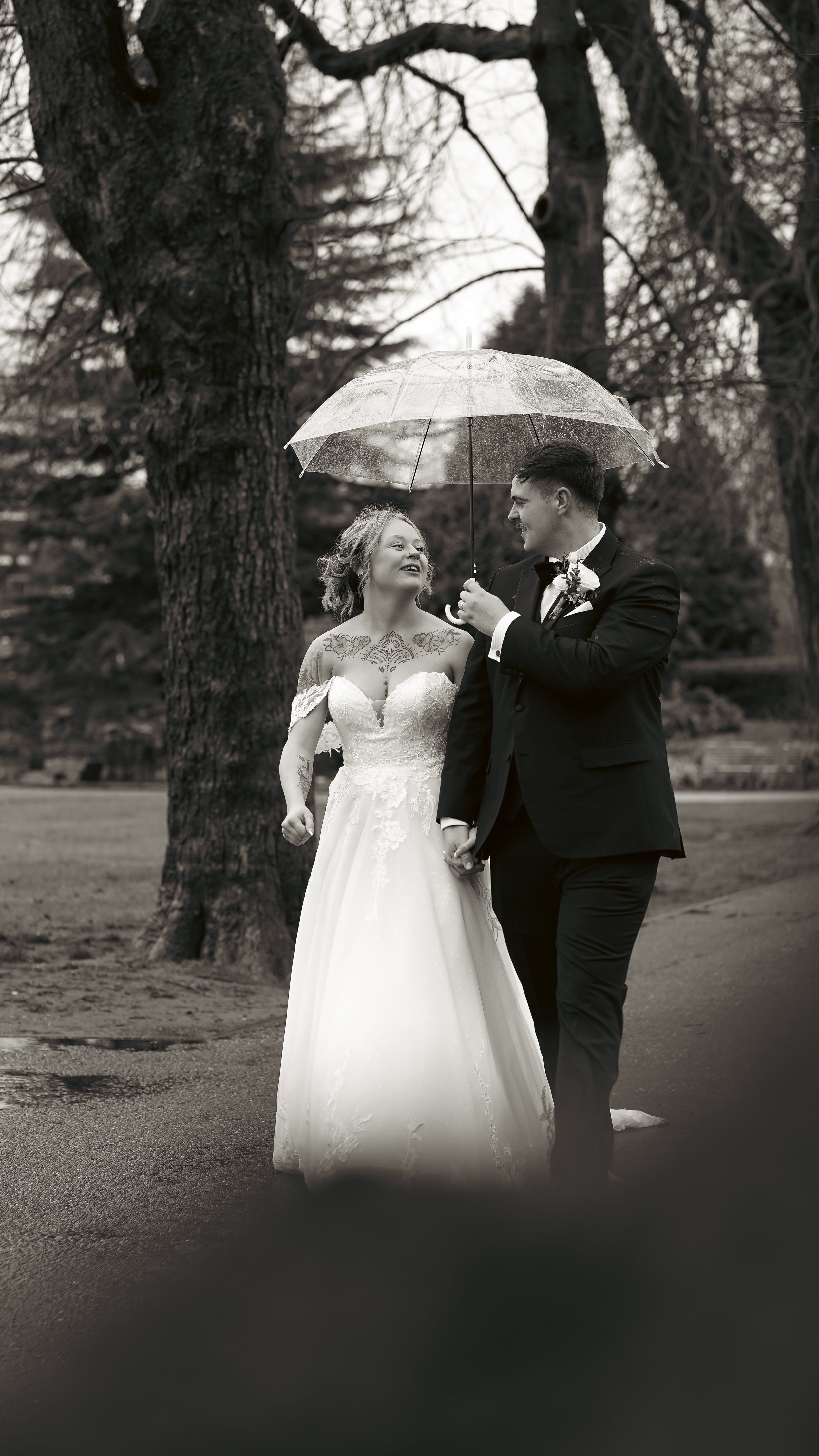 A newlywed couple walking outdoors on a rainy day, sharing a transparent umbrella. The bride is smiling and looking at the groom, who is holding her hand and the umbrella, wearing a black tuxedo. The bride is wearing a white wedding gown with lace de
