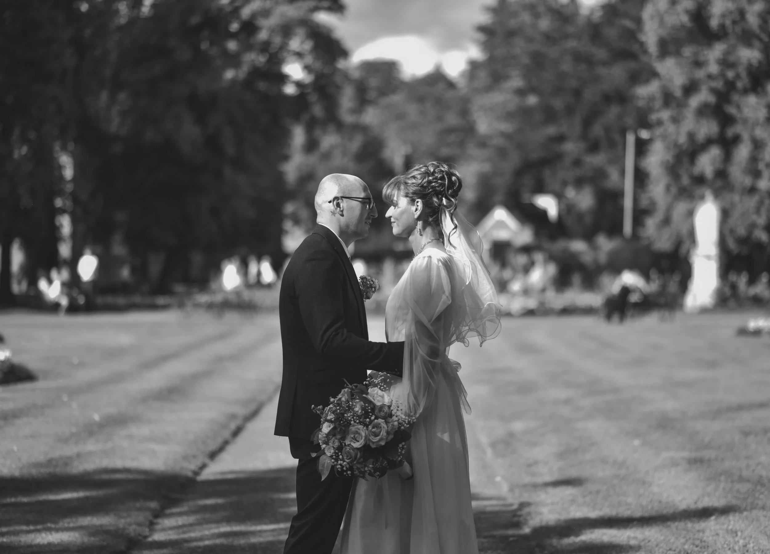 A black and white photo of a bride and groom standing face to face outdoors on a grassy area, holding hands, with trees and a house in the background.