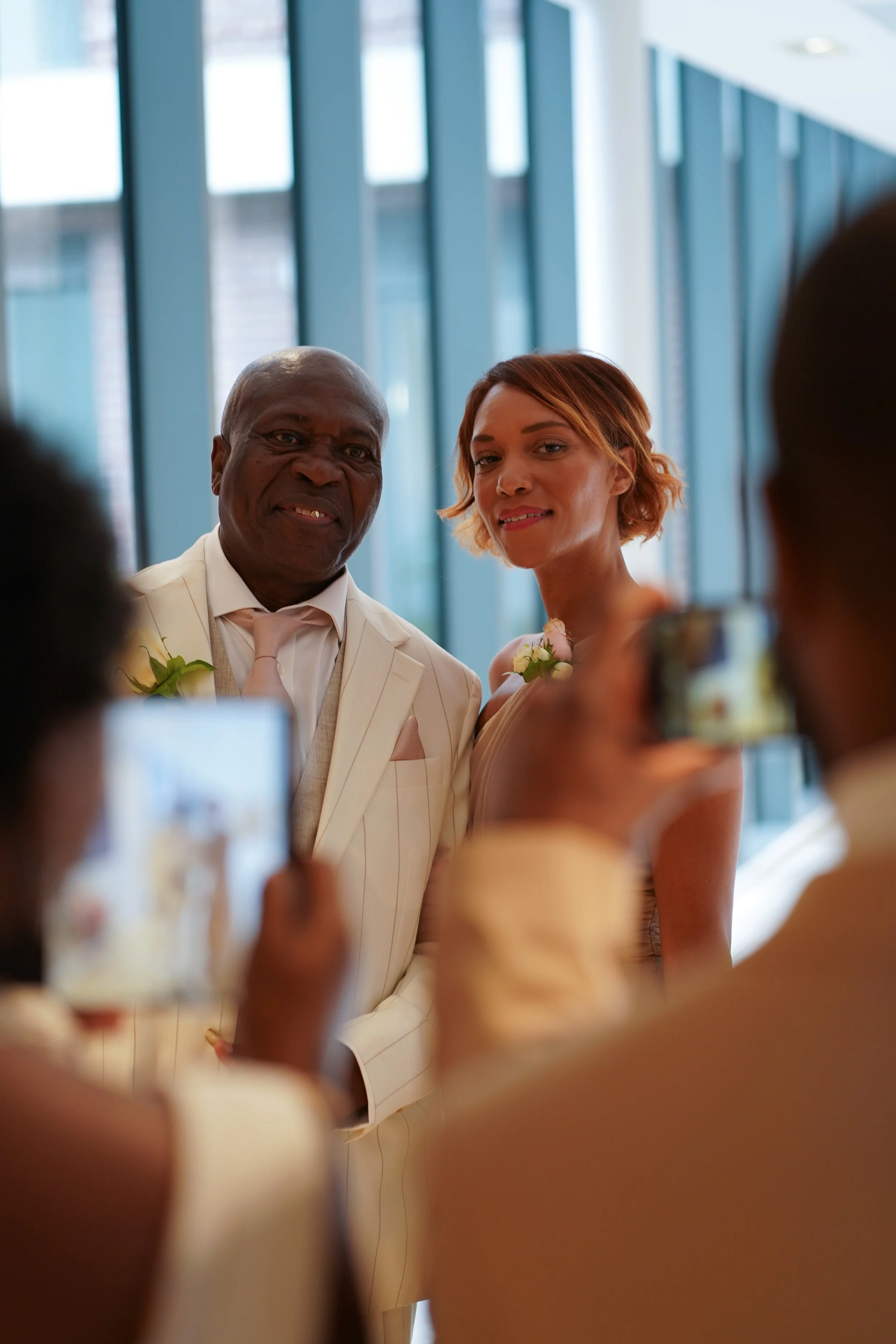 A man in a cream-coloured suit and a woman in a light satin dress taking a photo with a smartphone. The man is smiling and the woman has a slight smile. They are at a wedding or formal event with a glass wall in the background.