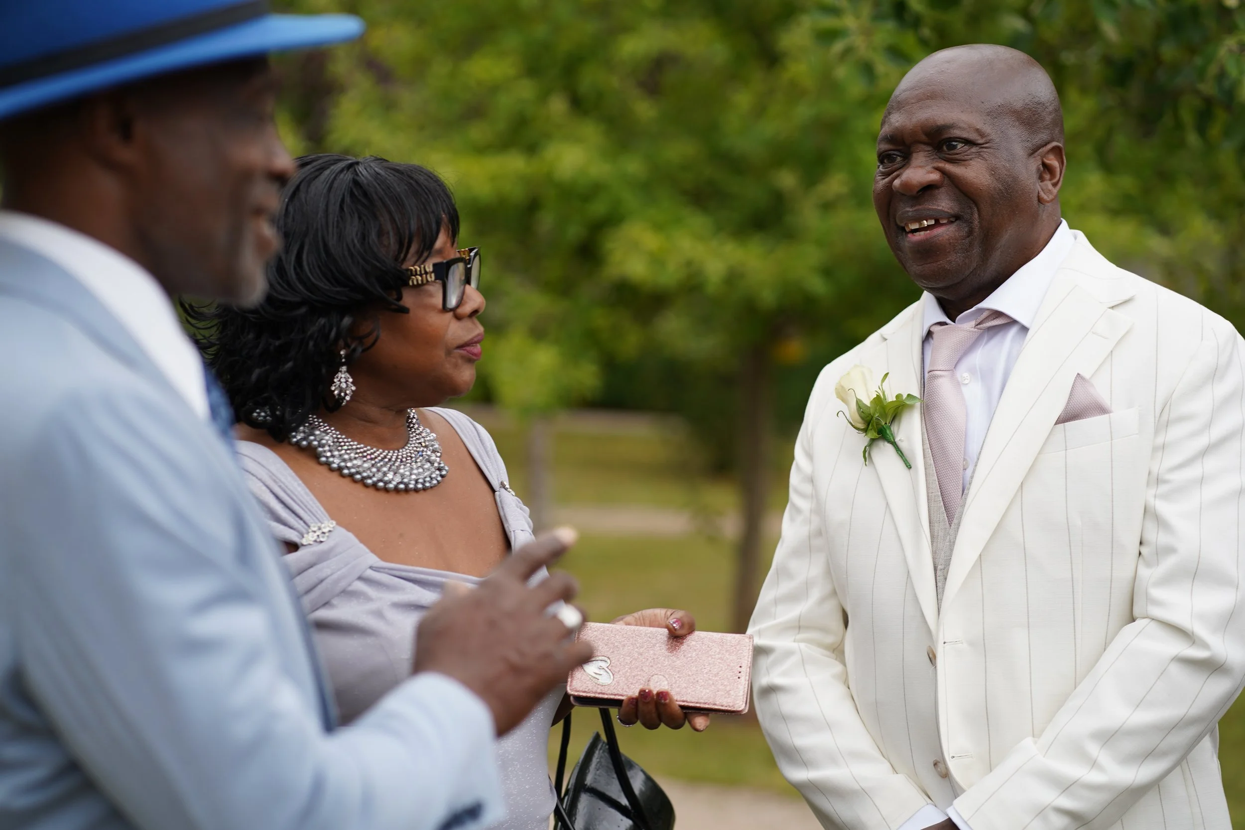 A wedding ceremony outdoors with a man in a white suit exchanging vows with a woman, while a man and woman stand nearby.