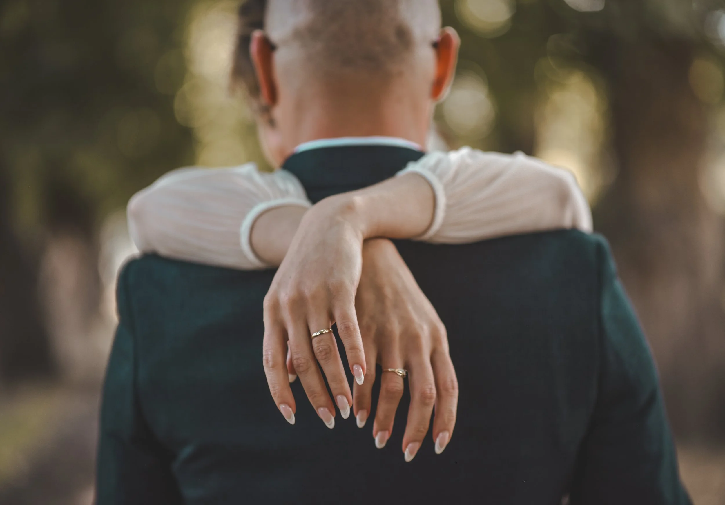 Close-up of a woman with manicured nails and rings on her fingers hugging a man in a dark suit outdoors with blurred trees in the background.