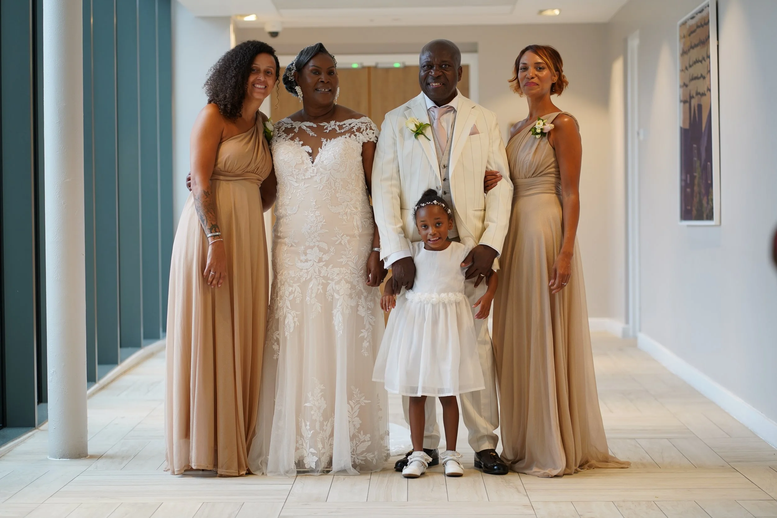 Group of six people, including a bride, groom, three women in similar dresses, and a young girl, standing in a hallway, dressed in formal wedding attire.