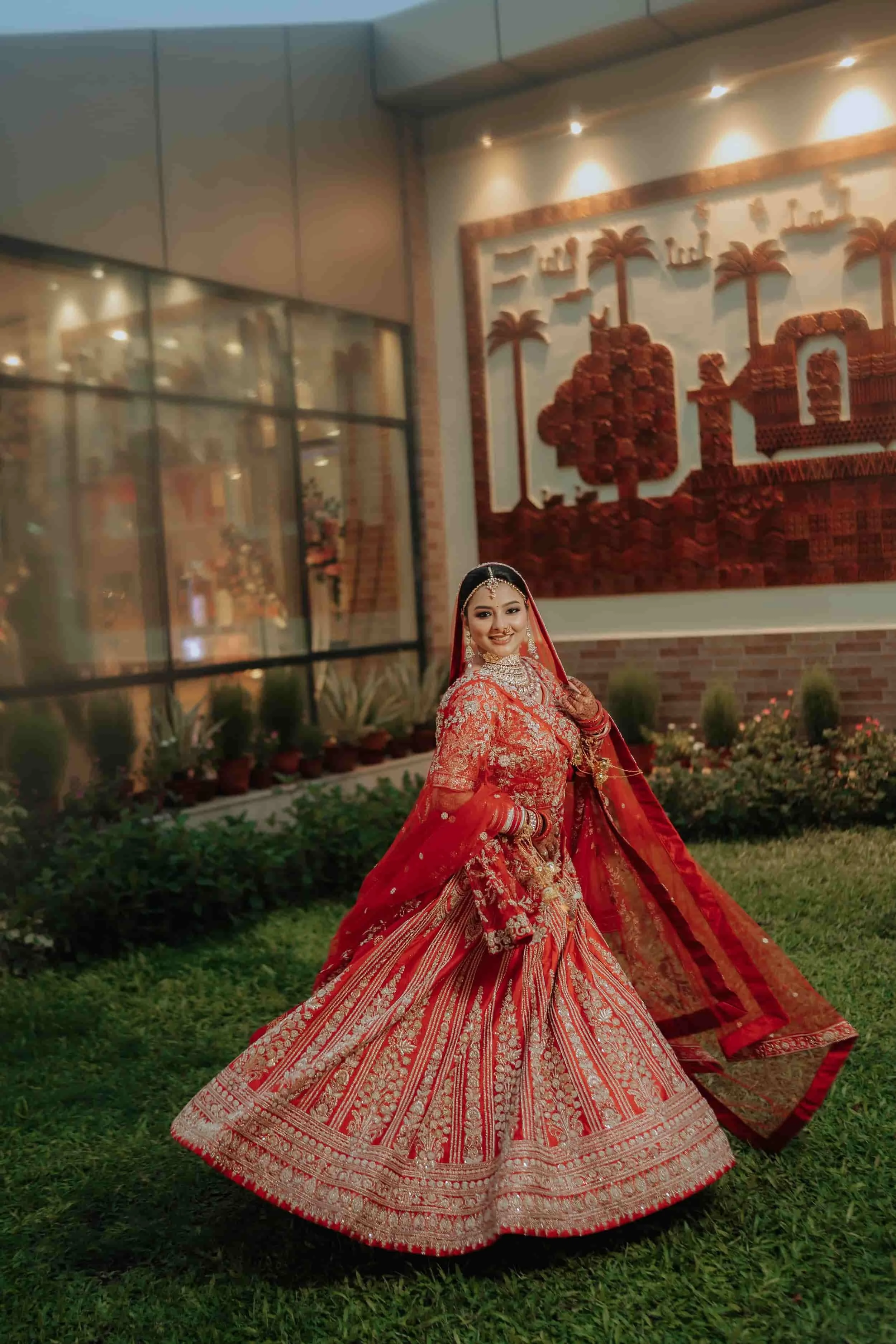 A woman dressed in a traditional red and gold bridal lehenga, smiling, standing outdoors on grass in front of a building with decorative wall art and plants.