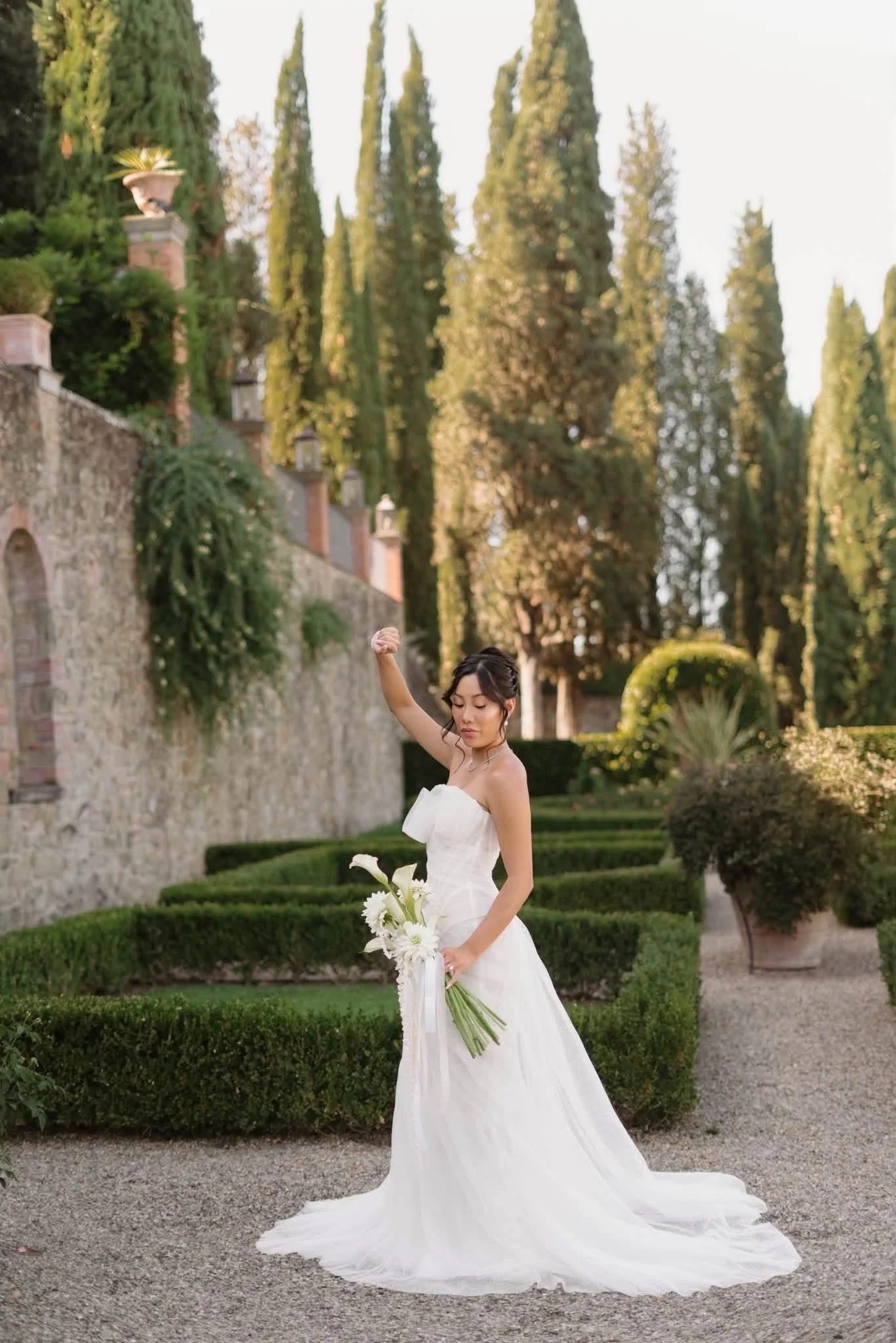 A bride in a strapless wedding gown holding a bouquet of white flowers stands in a garden with neatly trimmed hedges, stone walls, and tall cypress trees in the background.