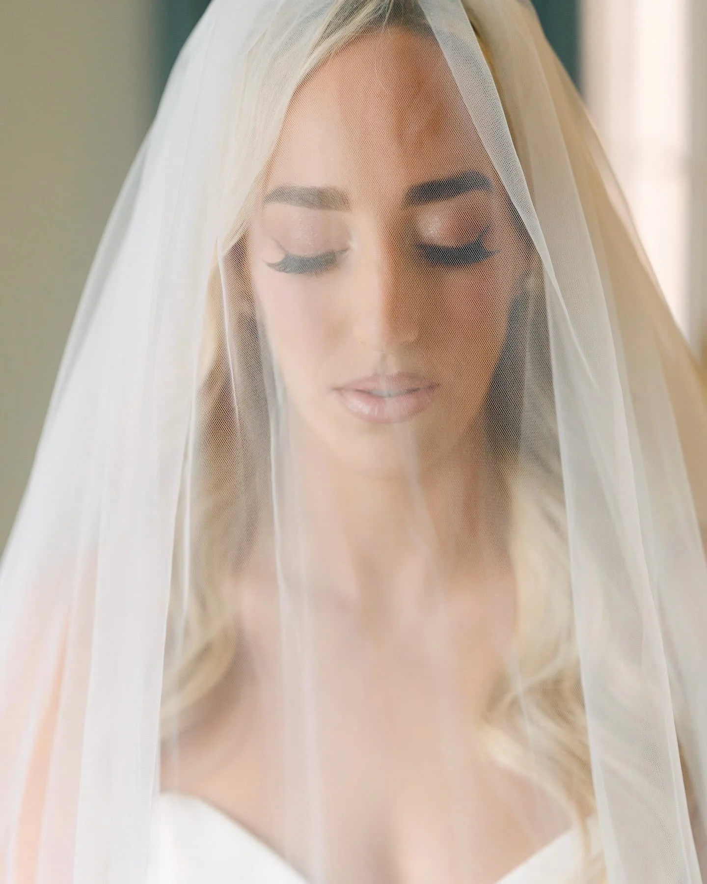 A woman with blonde hair, closed eyes, and makeup, wearing a white veil over her face, with a serene expression.