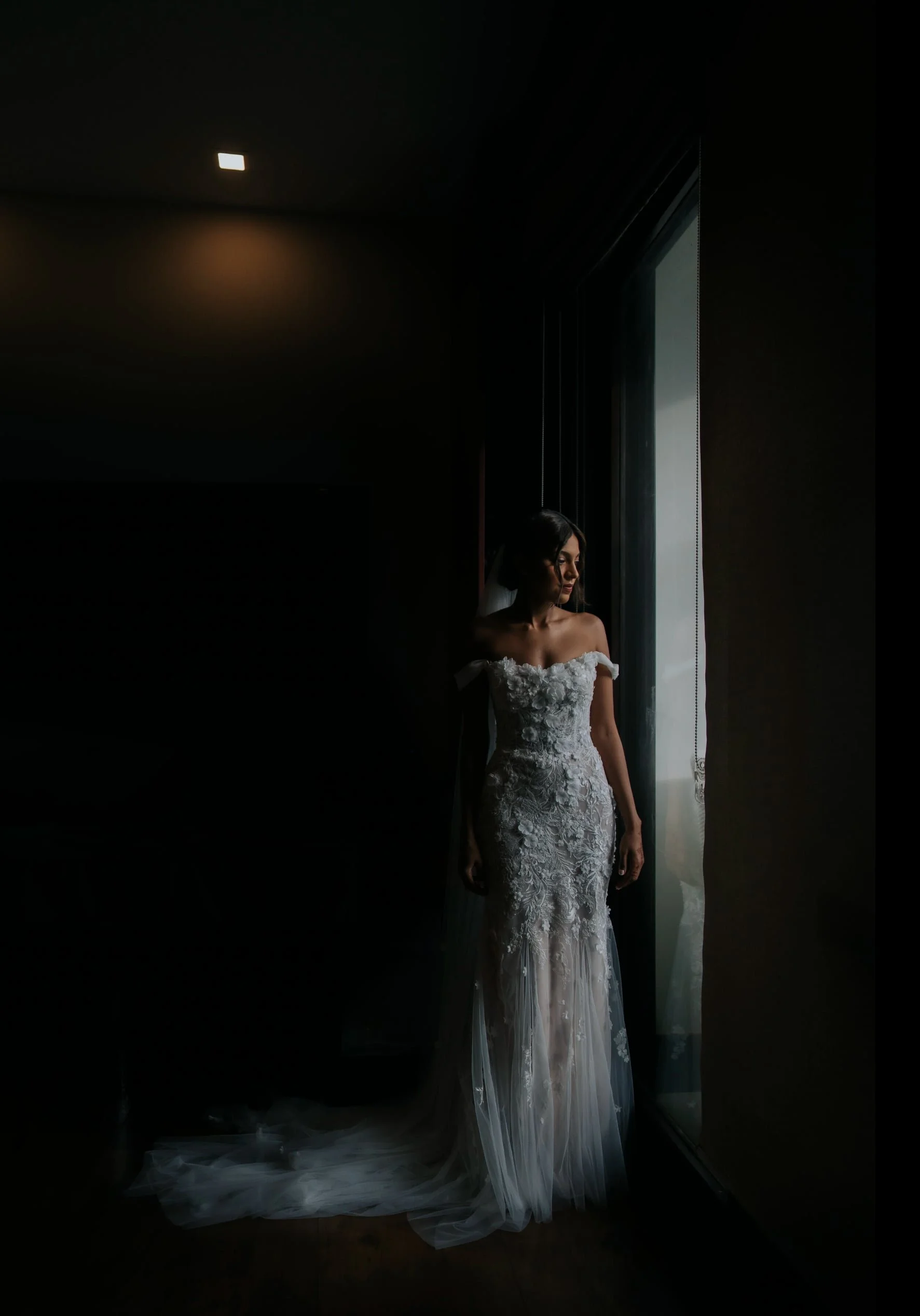 A woman in a white wedding dress standing by a window, with light illuminating her face and dress, against a dark interior background.