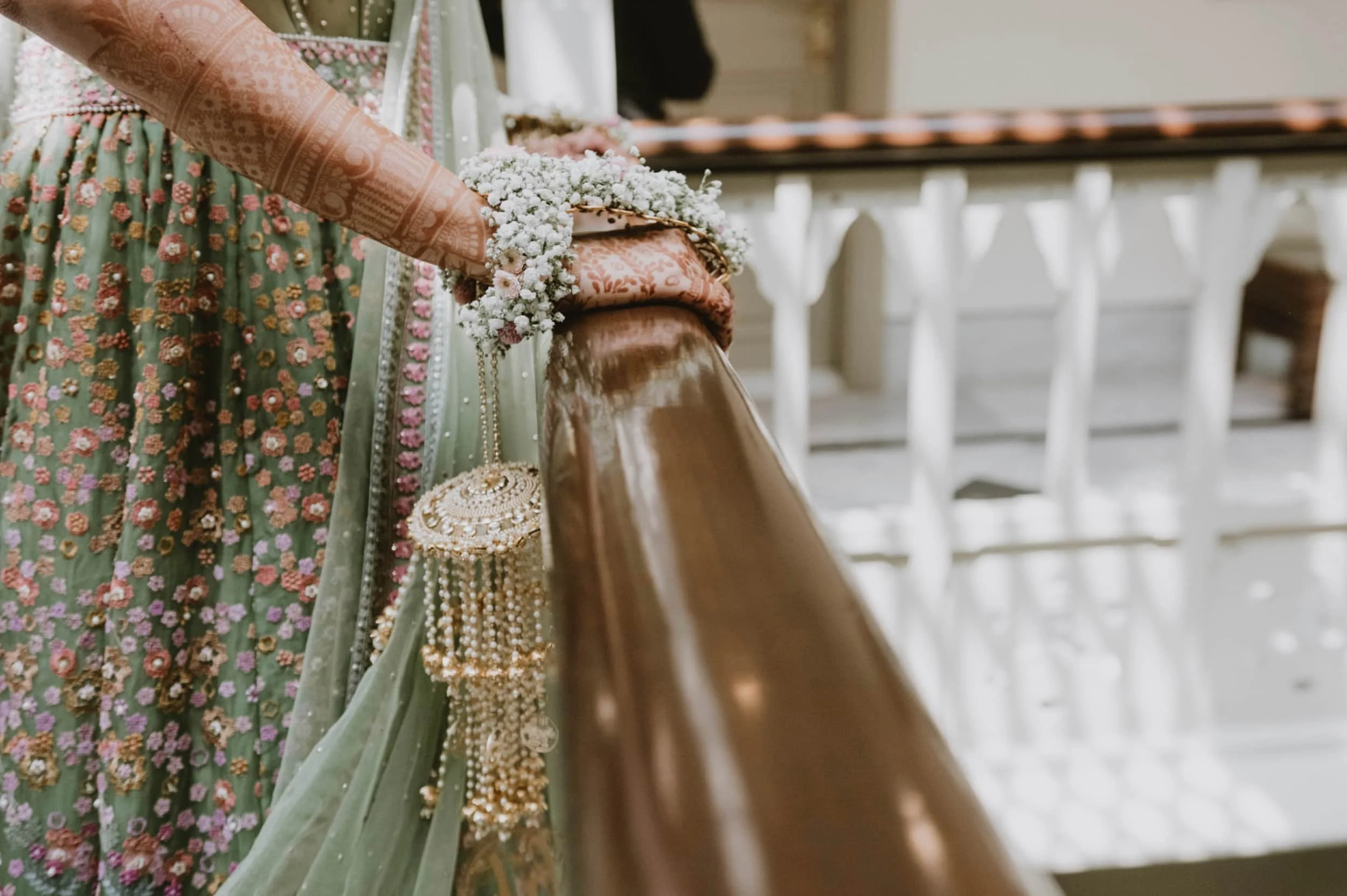 Close-up of an ornate pink and peach wedding dress with floral embroidery and beadwork. A bouquet of white flowers rests on a folded fabric, with a pearl necklace hanging from it. A wooden pew and white architectural details are visible in the backgr