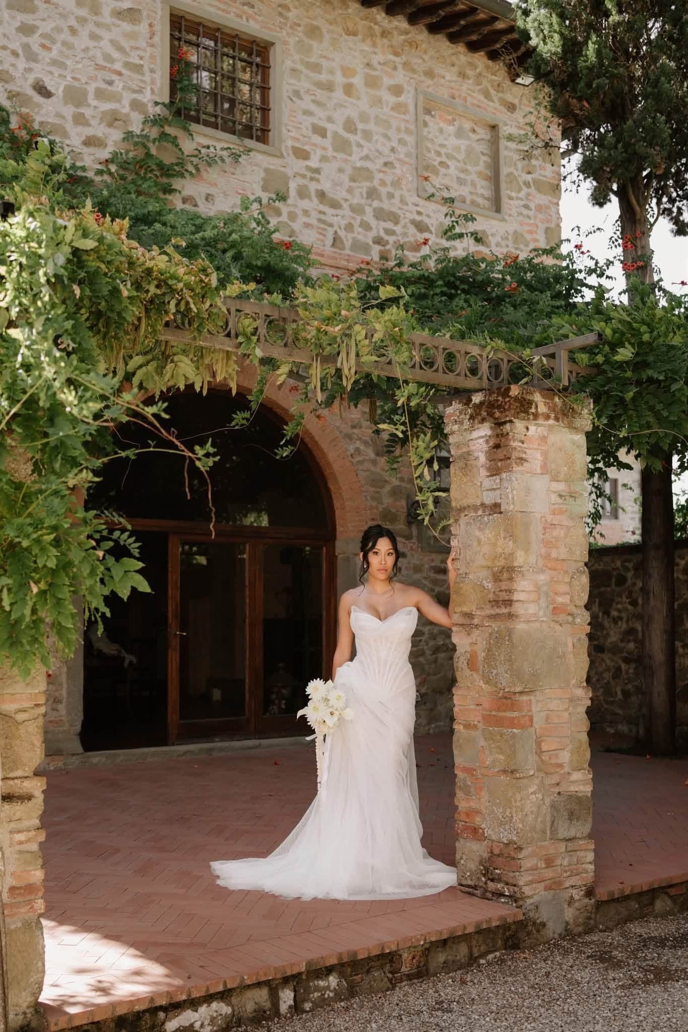 A bride in a white wedding gown holding a bouquet, standing under an arched brick and stone structure with greenery and a tree in the background.