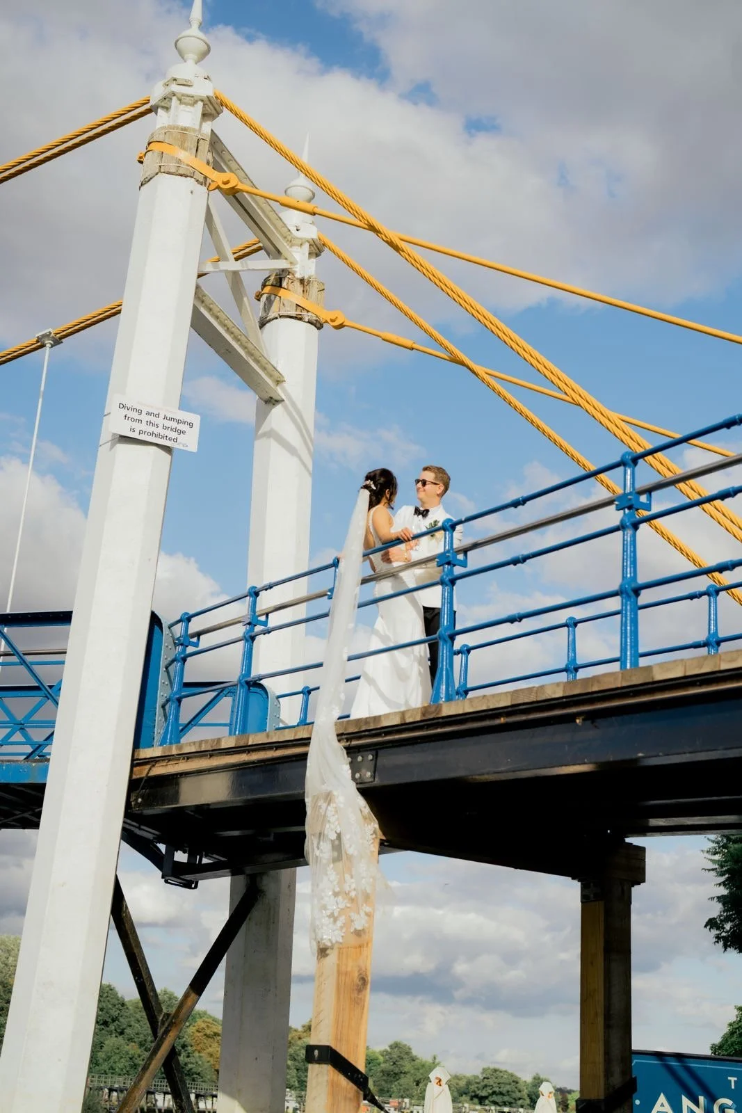 A bride and groom are dancing on a bridge with a cloudy sky in the background.