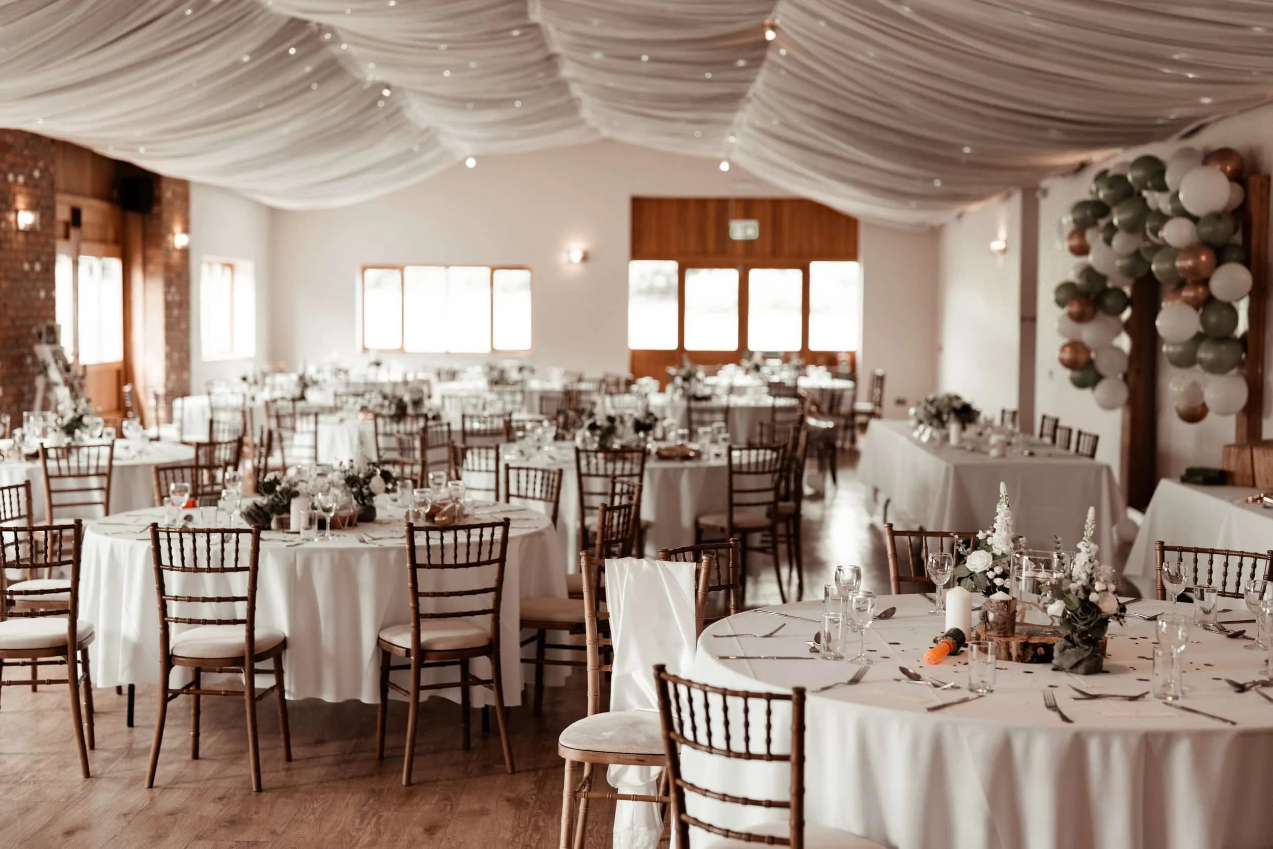 A decorated banquet hall with round tables covered with white tablecloths, elegant chairs, and floral centerpieces. The ceiling features draped fabric with small twinkle lights. Balloons are arranged on the wall, and large windows let in natural ligh