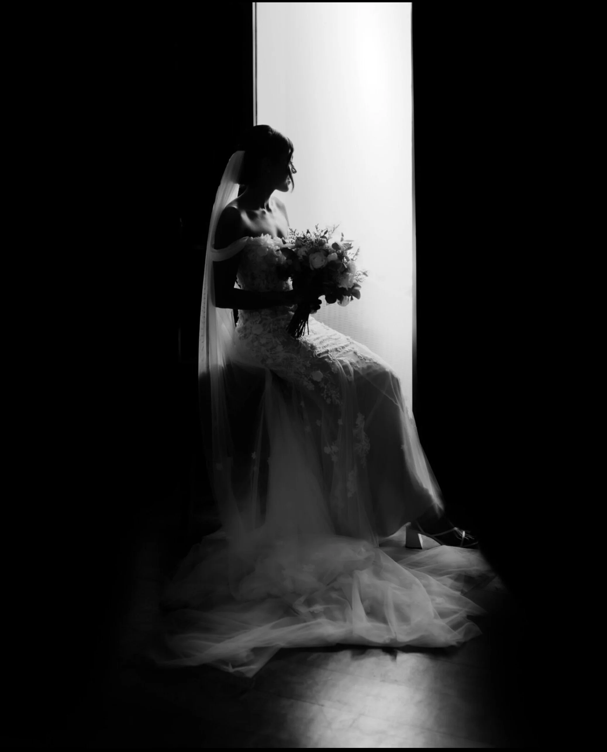 Silhouette of a bride sitting in front of a large illuminated window, holding a bouquet of flowers, wearing a wedding dress and veil, with long tulle fabric flowing around her on the floor.