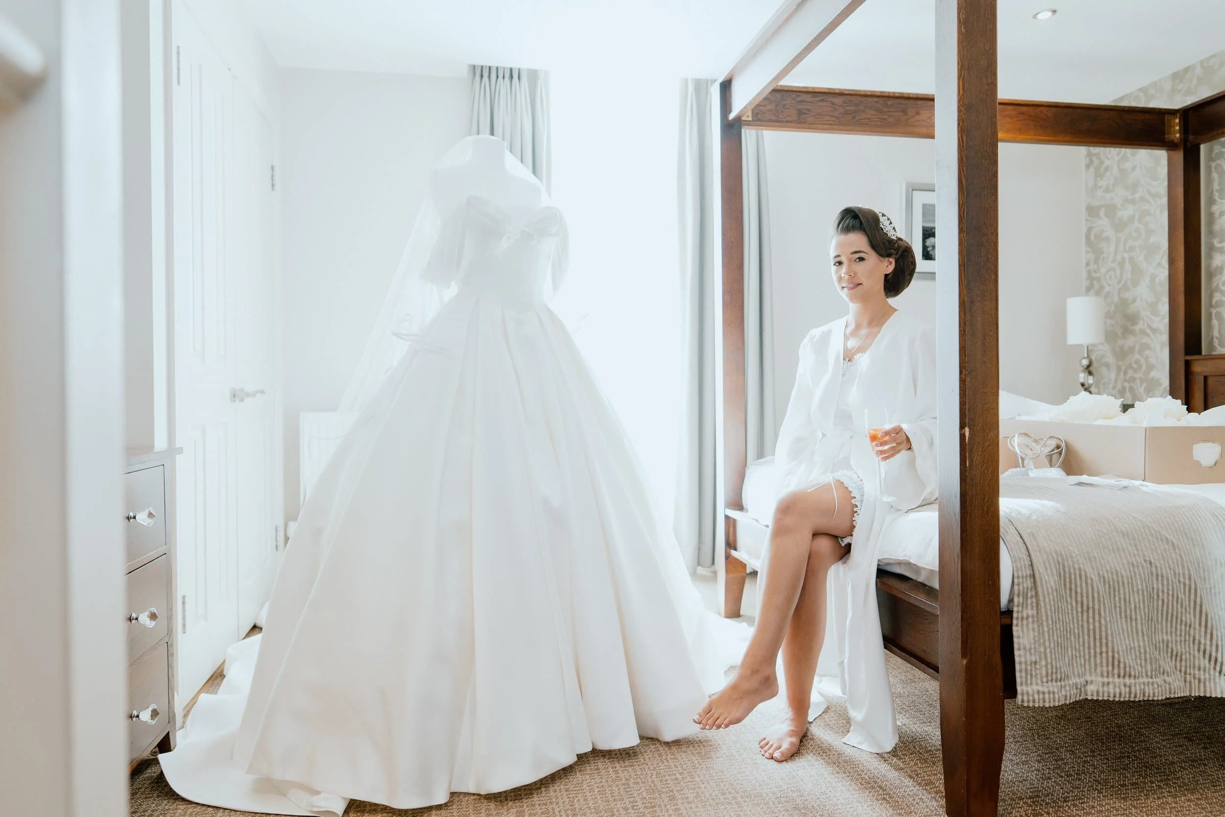 A woman sitting on a bed in a hotel room, wearing a white robe, holding a glass of orange juice, and looking into a mirror with a wedding dress on it.