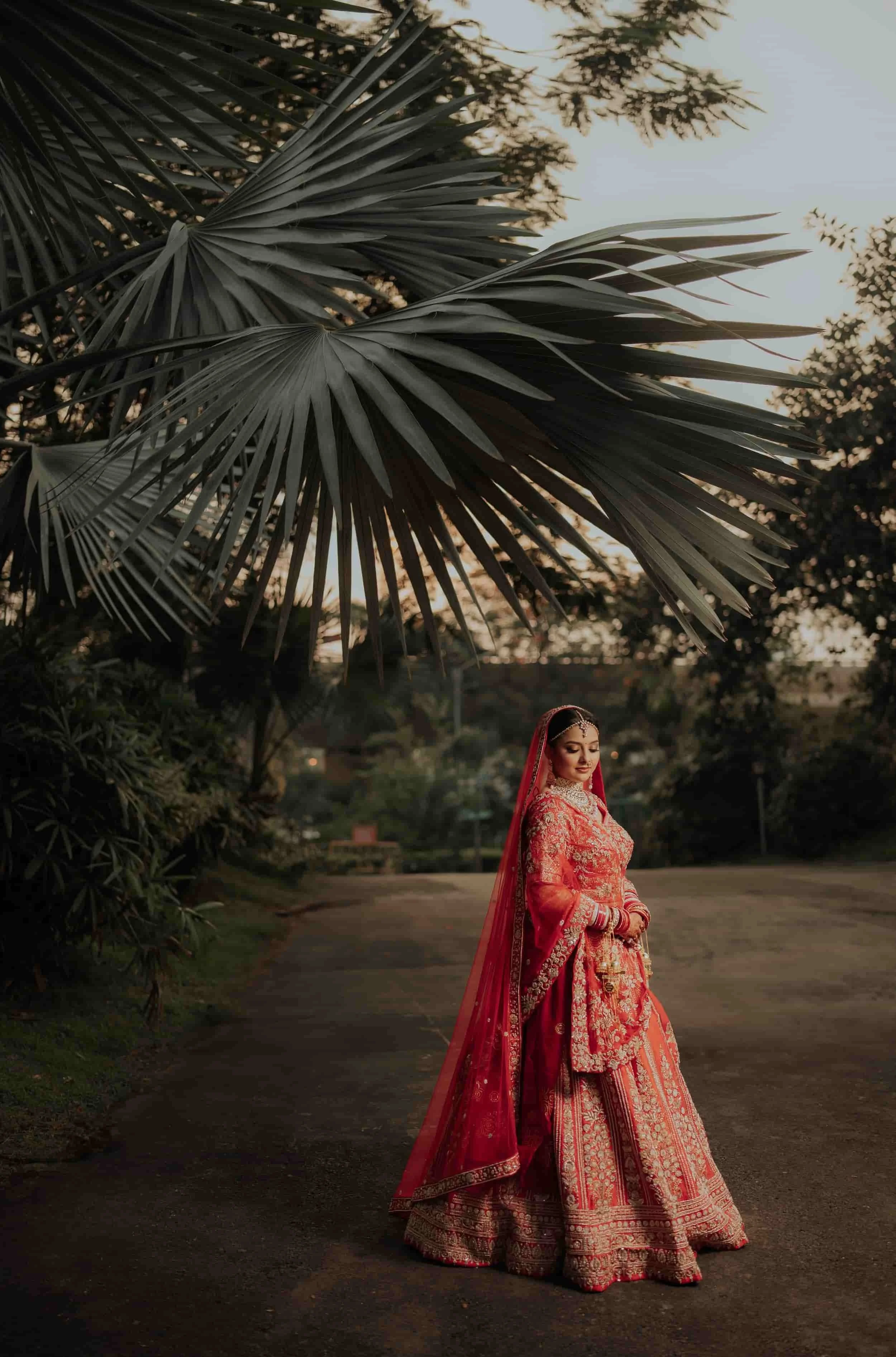 A woman dressed in a traditional red and gold ornate bridal outfit with intricate embroidery, standing outdoors during sunset or early evening, with large palm leaves and trees in the background.