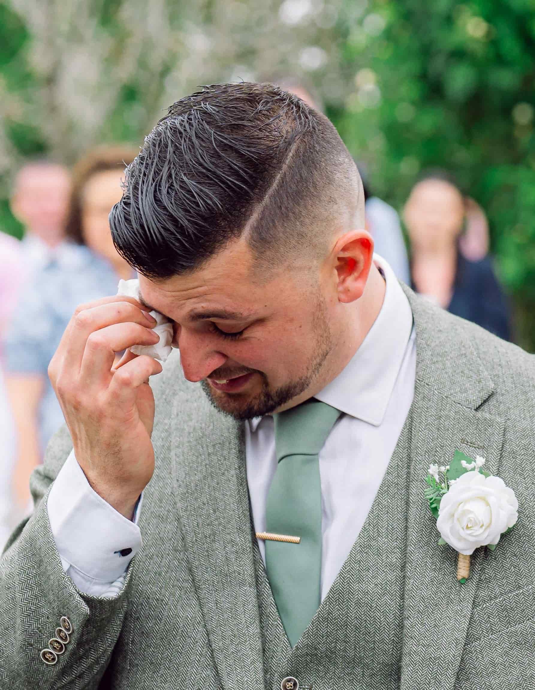 A close-up of a man in a grey checked suit with a white shirt and light green tie, wiping his eye with a tissue, during a wedding or formal event. He has a boutonniere of white roses and greenery on his suit lapel. The background features blurred peo