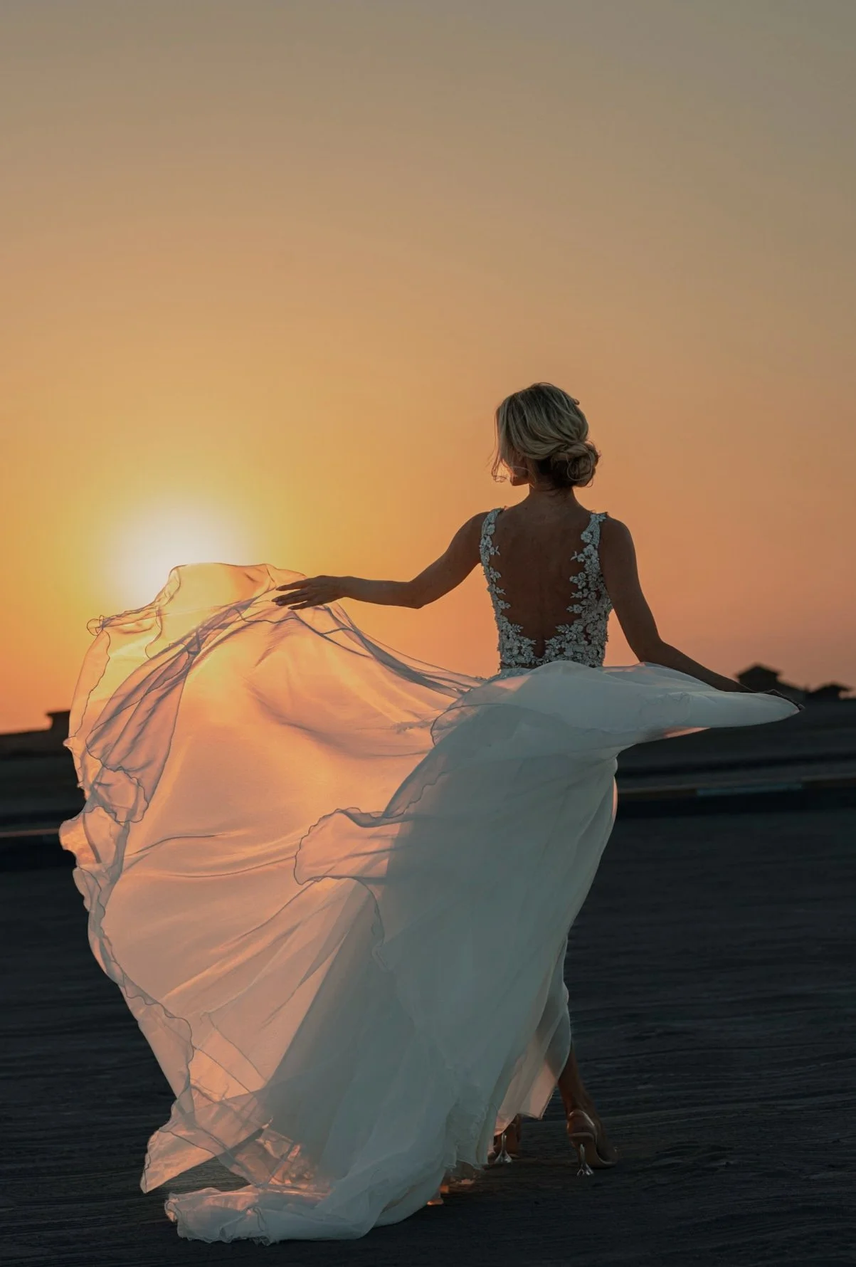 A woman in a wedding dress, standing outdoors during sunset, with her back to the camera, holding the flowing fabric of her dress.