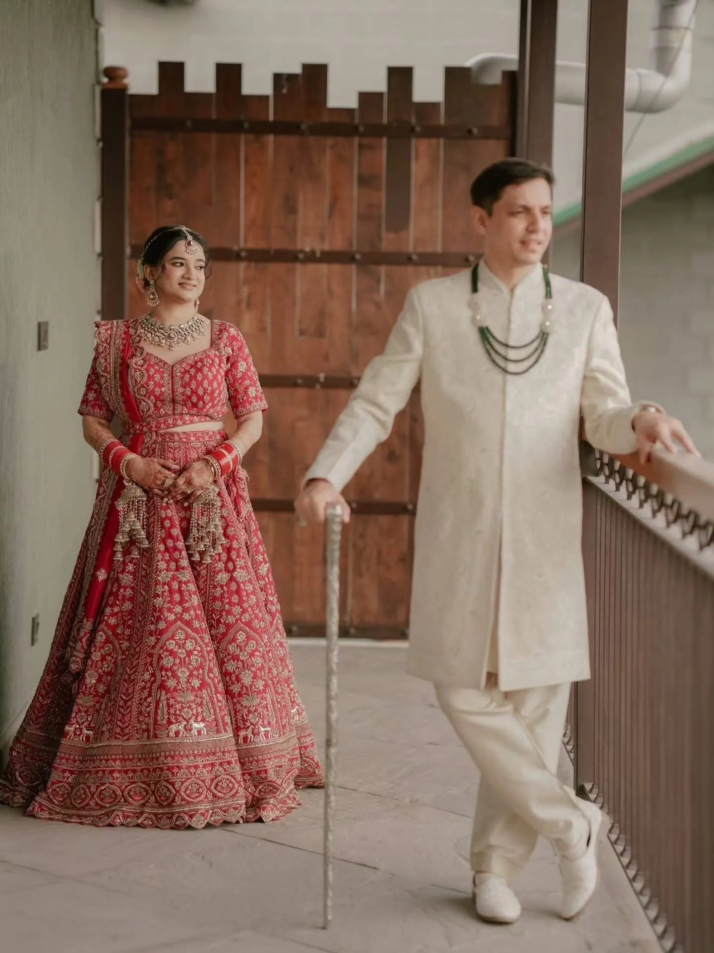 A woman dressed in traditional Indian bridal attire and jewelry stands beside a man dressed in traditional Indian cream-colored attire with necklaces, as he leans on a railing.