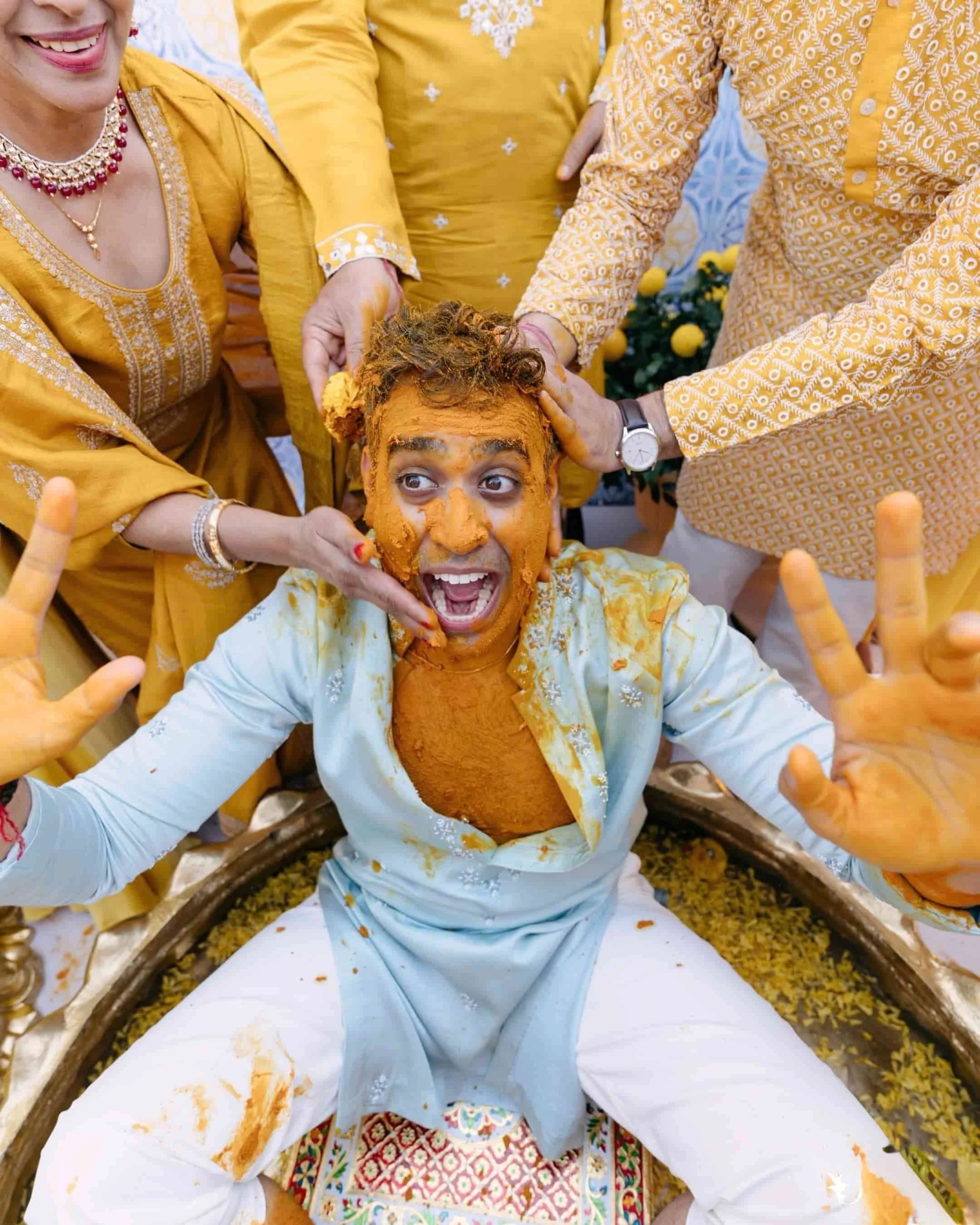 A person participating in a traditional Indian wedding ceremony, with yellow-orange turmeric paste applied to their face and body, surrounded by others in yellow and white attire.