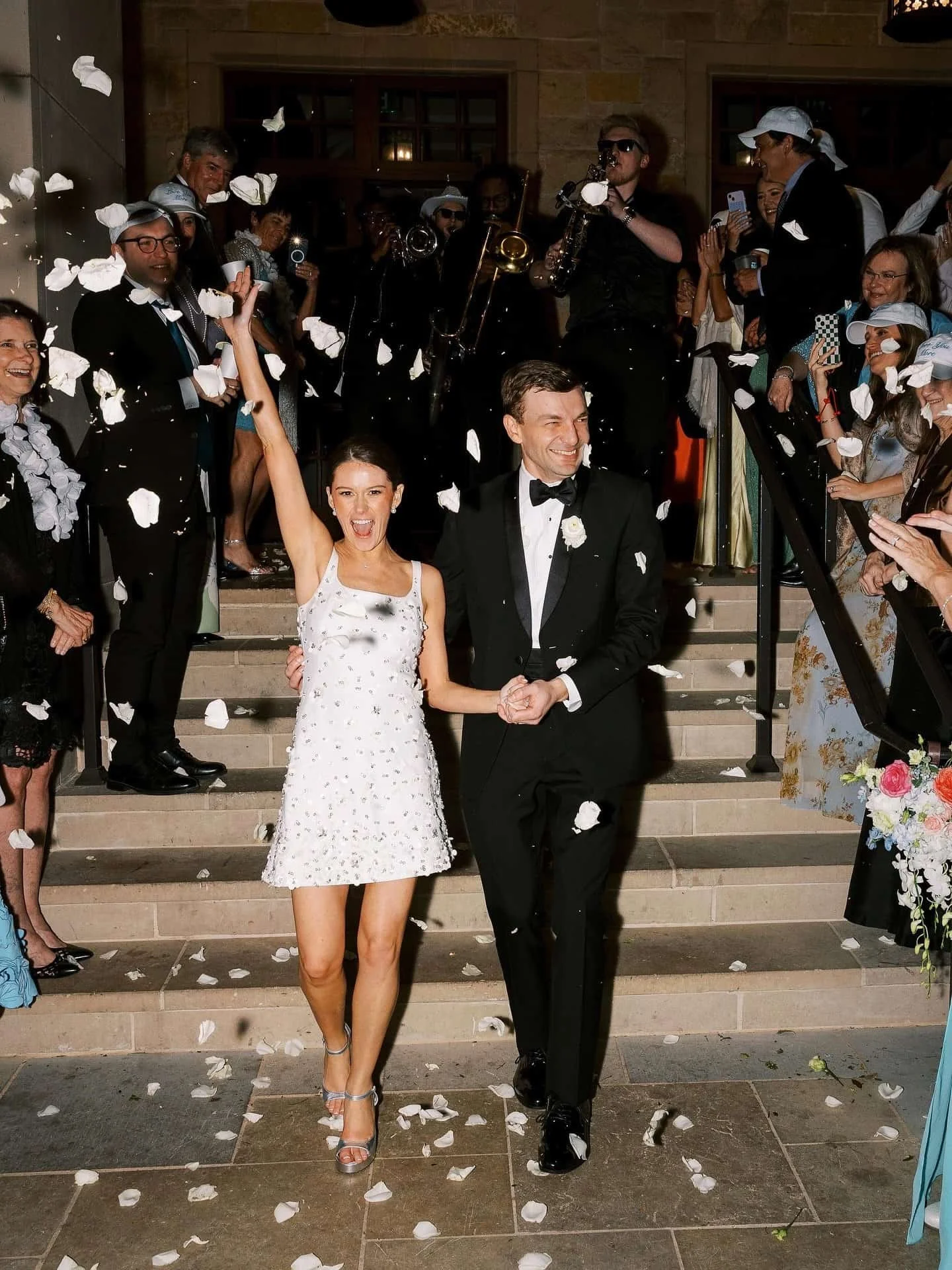 A bride and groom walking down stairs at their wedding reception, surrounded by guests throwing flower petals. The bride is in a white dress and the groom is in a tuxedo.