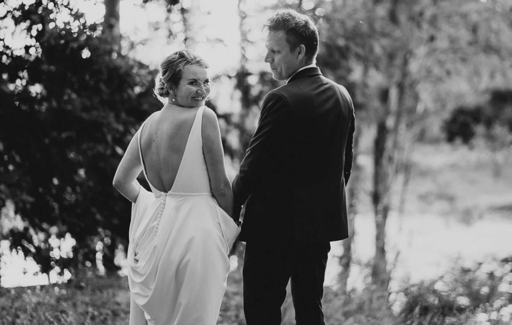 A bride and groom standing outdoors, facing each other, with the bride smiling and the groom looking at her. The bride is wearing a sleeveless wedding gown and the groom is dressed in a dark suit. Trees and sunlight create a blurred, natural backgrou