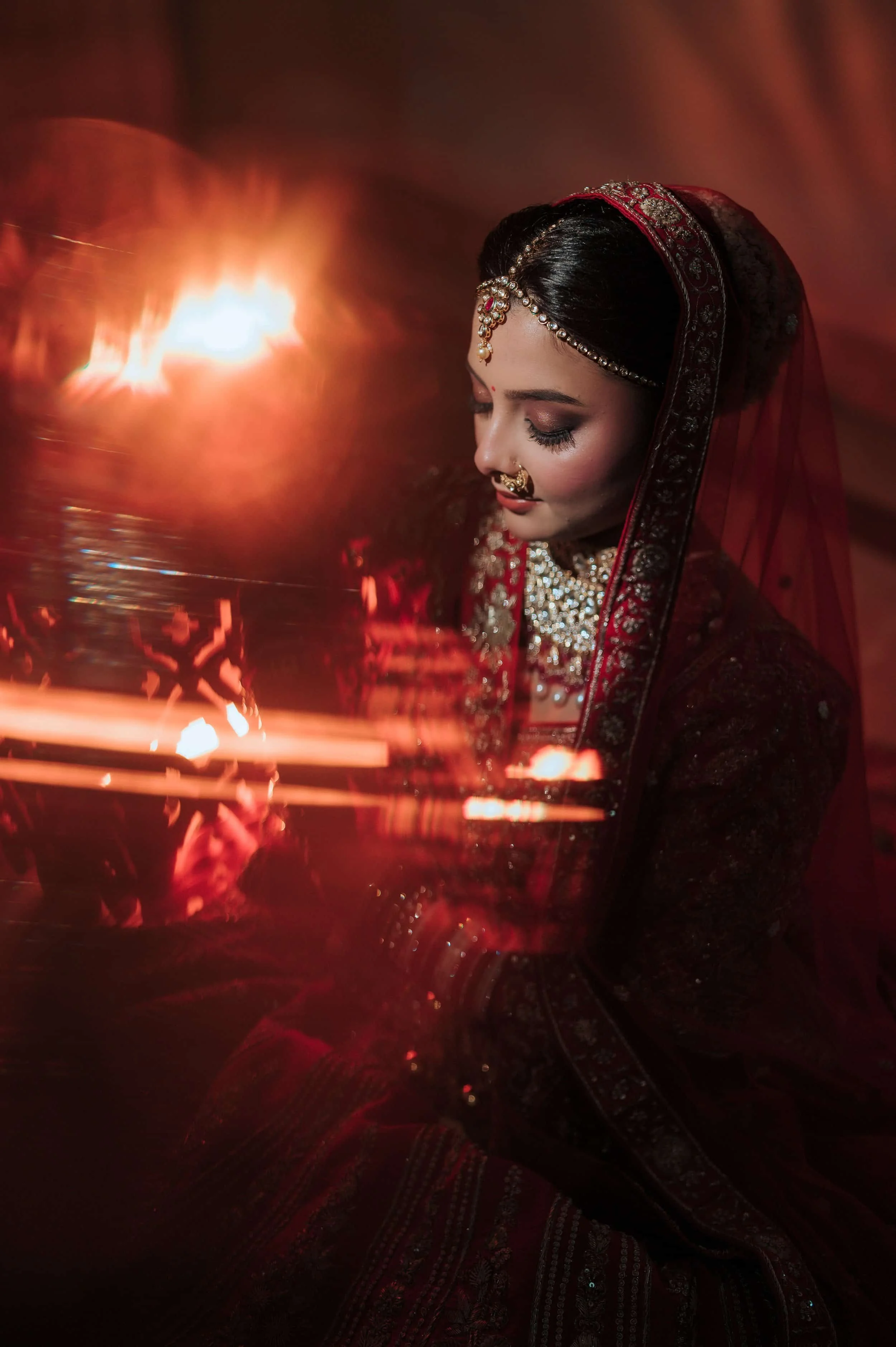 A woman dressed in traditional Indian bridal attire with jewelry, looking down with closed eyes, illuminated by soft lighting and lanterns, with red and gold adornments.