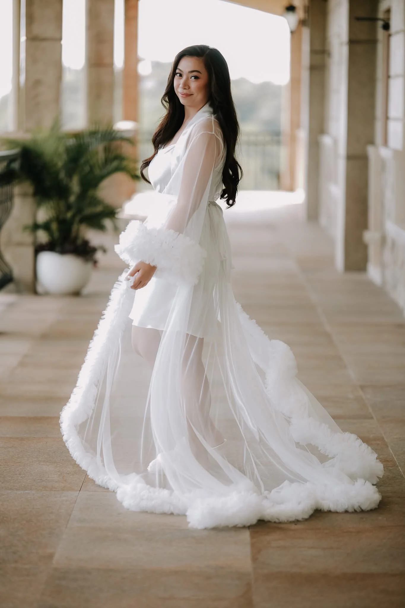A woman with long dark hair in a white wedding dress standing in a bright hallway with large windows and wooden flooring, holding her train.