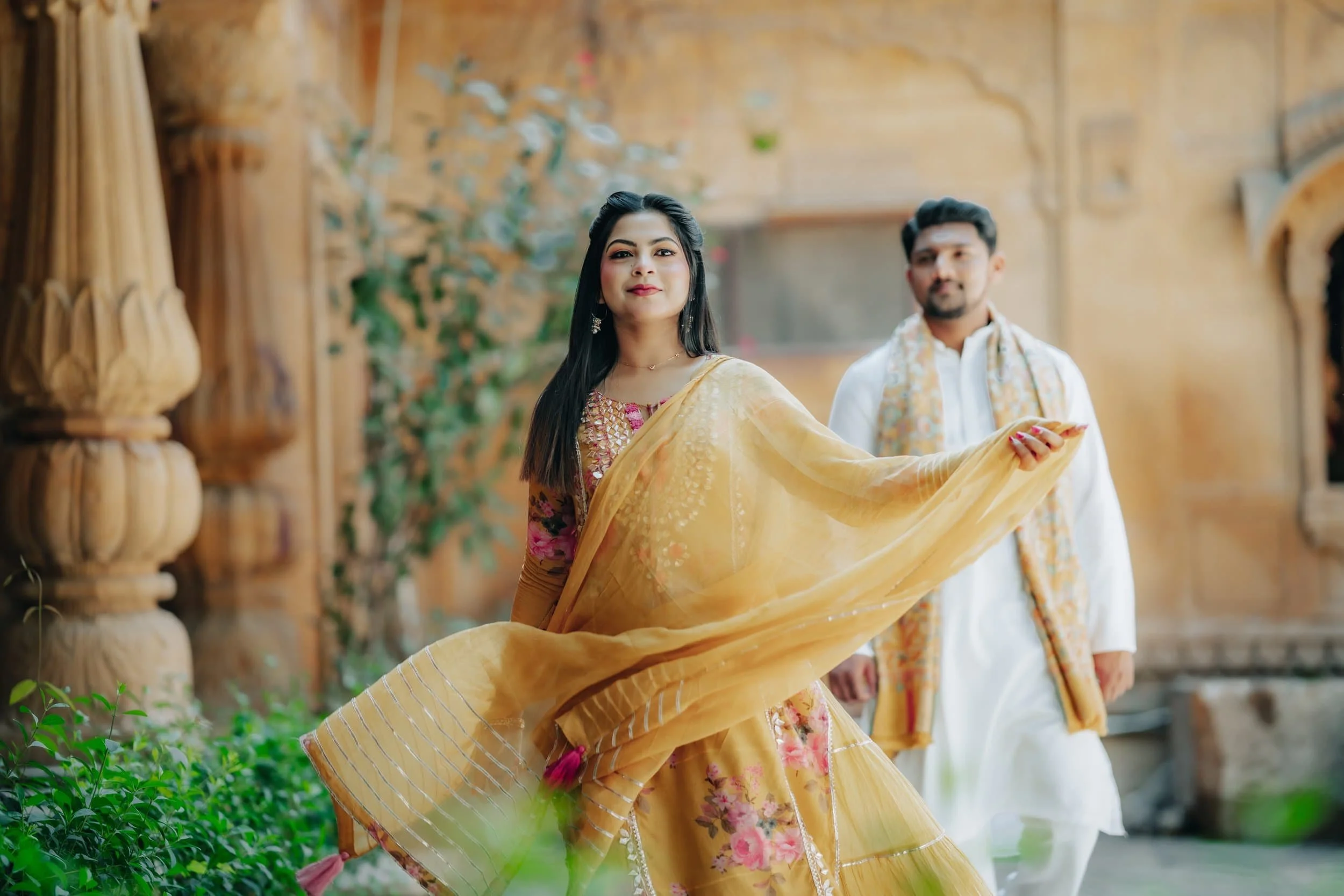 An Indian woman in traditional attire holding a yellow dupatta, standing outdoors with a man in similar traditional clothing in the background, amidst a decorative wooden structure and greenery.