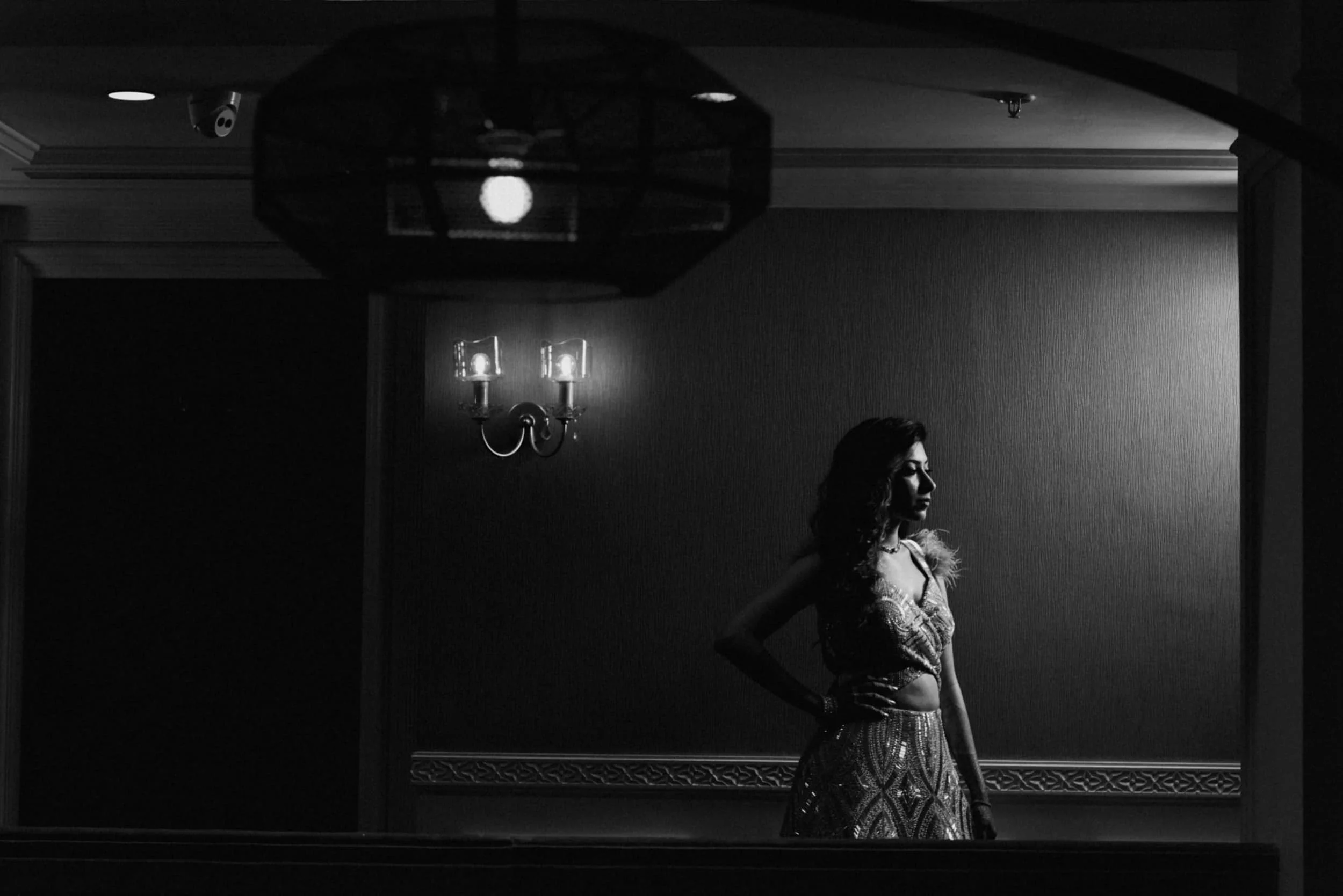 Black and white photograph of a woman standing sideways in a room with minimal decor, a wall sconce, and a ceiling light fixture.