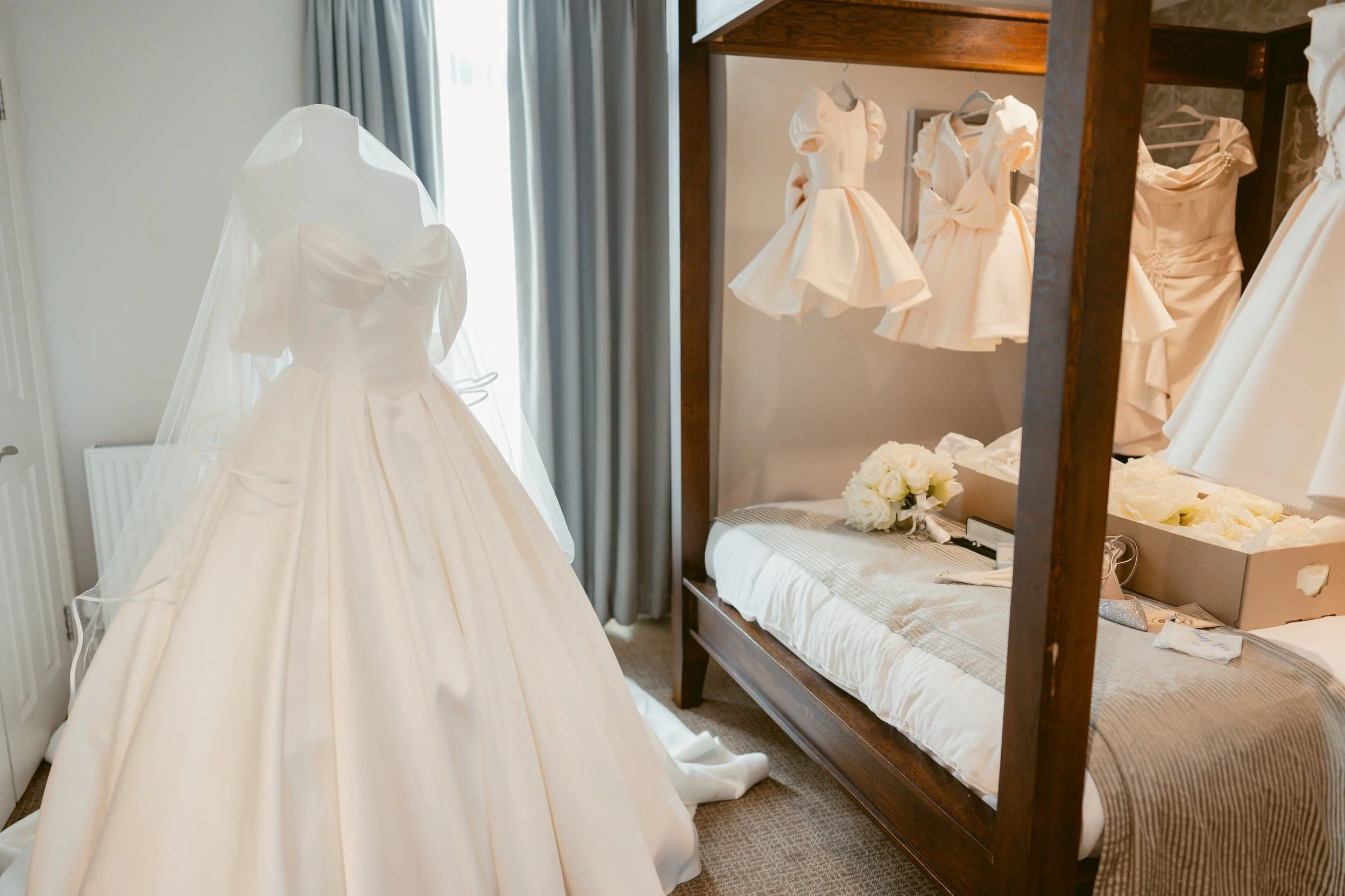 Wedding dresses displayed in a room, with a bed and a wooden shelving unit.