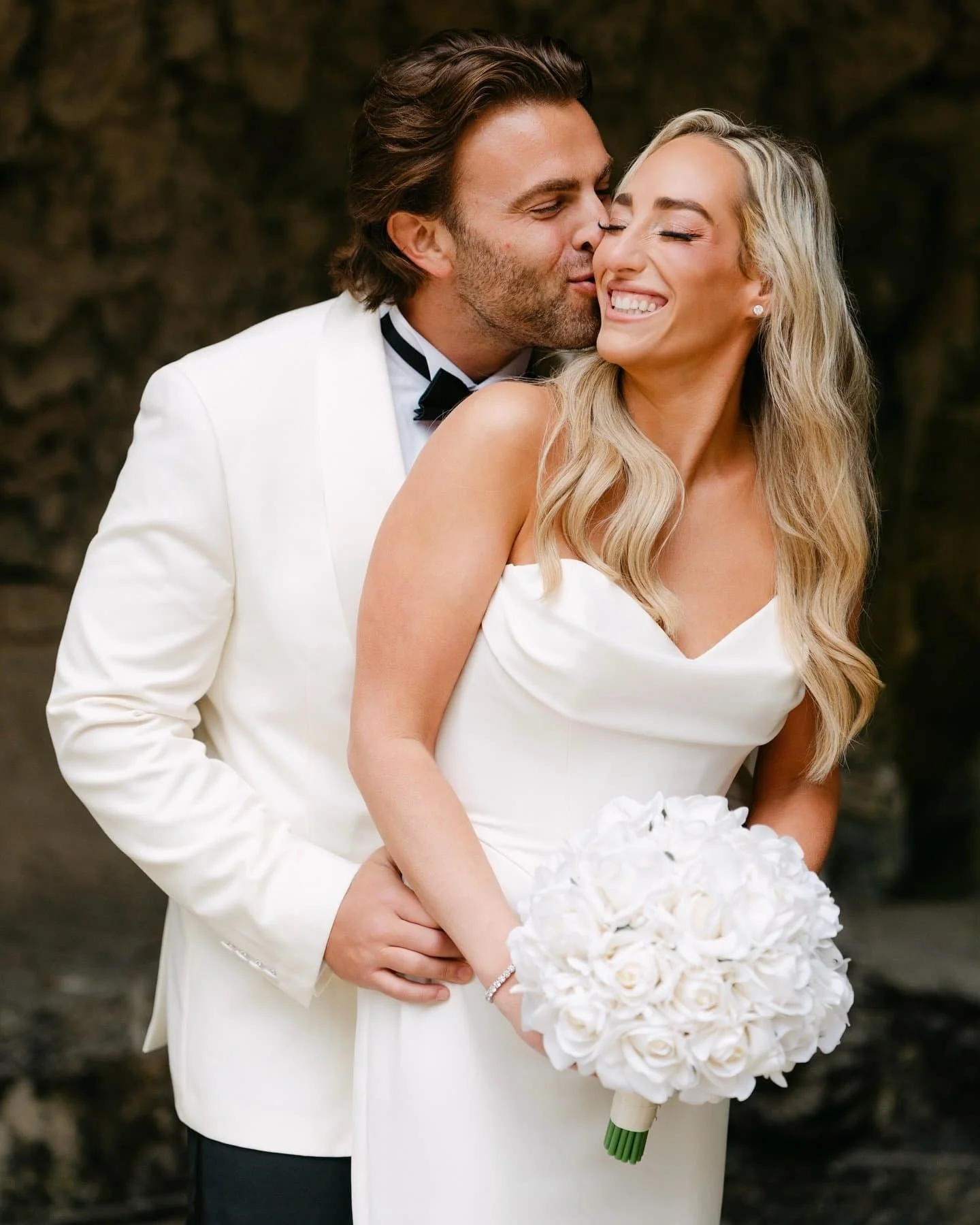 A newlywed couple in wedding attire, with the groom kissing the bride on the cheek while she smiles with eyes closed, holding a bouquet of white roses, against a dark natural background.