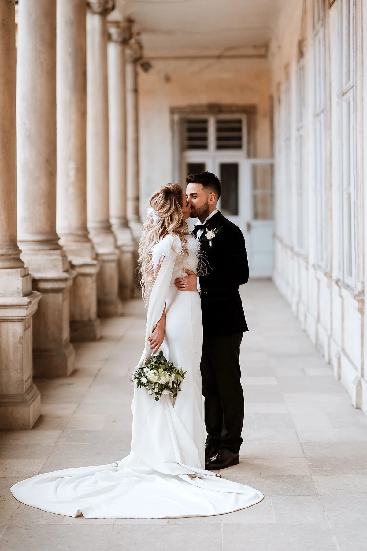 A bride and groom kiss in a historic corridor with stone columns and large windows. The bride is holding a bouquet, dressed in a white wedding gown with a long train, and the groom is in a black tuxedo with a boutonniere.