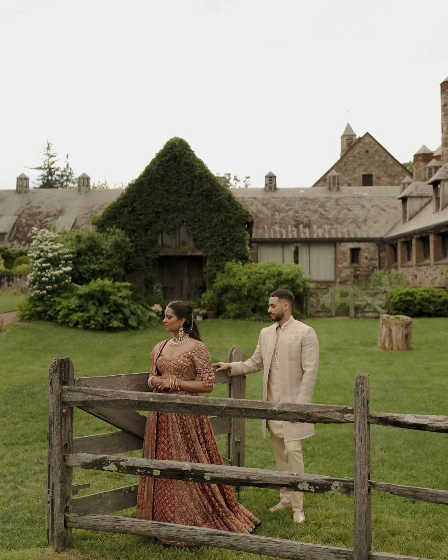 A man and woman dressed in traditional South Asian attire standing outdoors near a wooden fence with a historic stone building and lush greenery in the background.