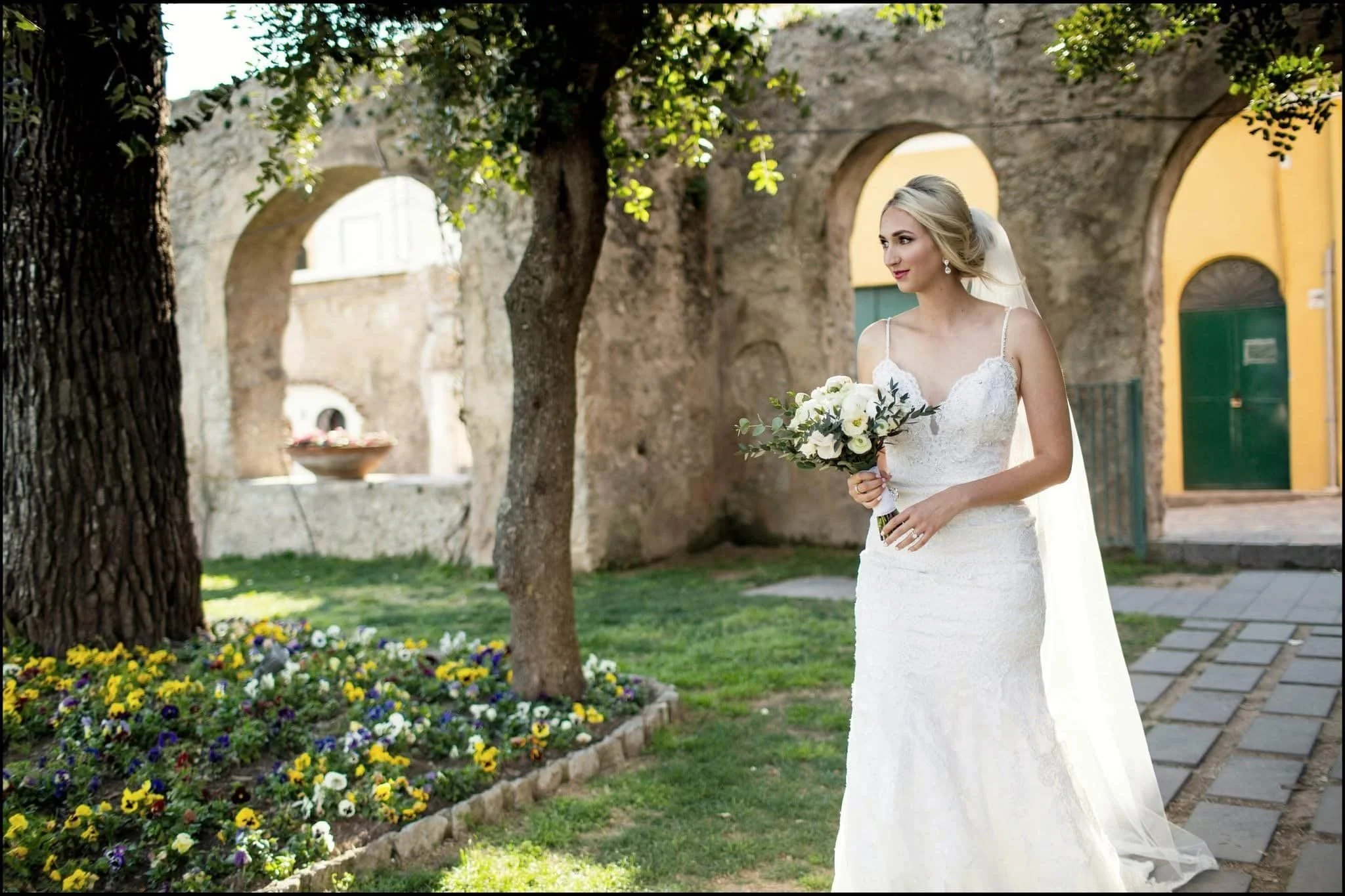 A bride in a white wedding dress holding a bouquet of white flowers, standing outdoors near a stone wall and colorful flower bed, with trees and yellow and green buildings in the background.