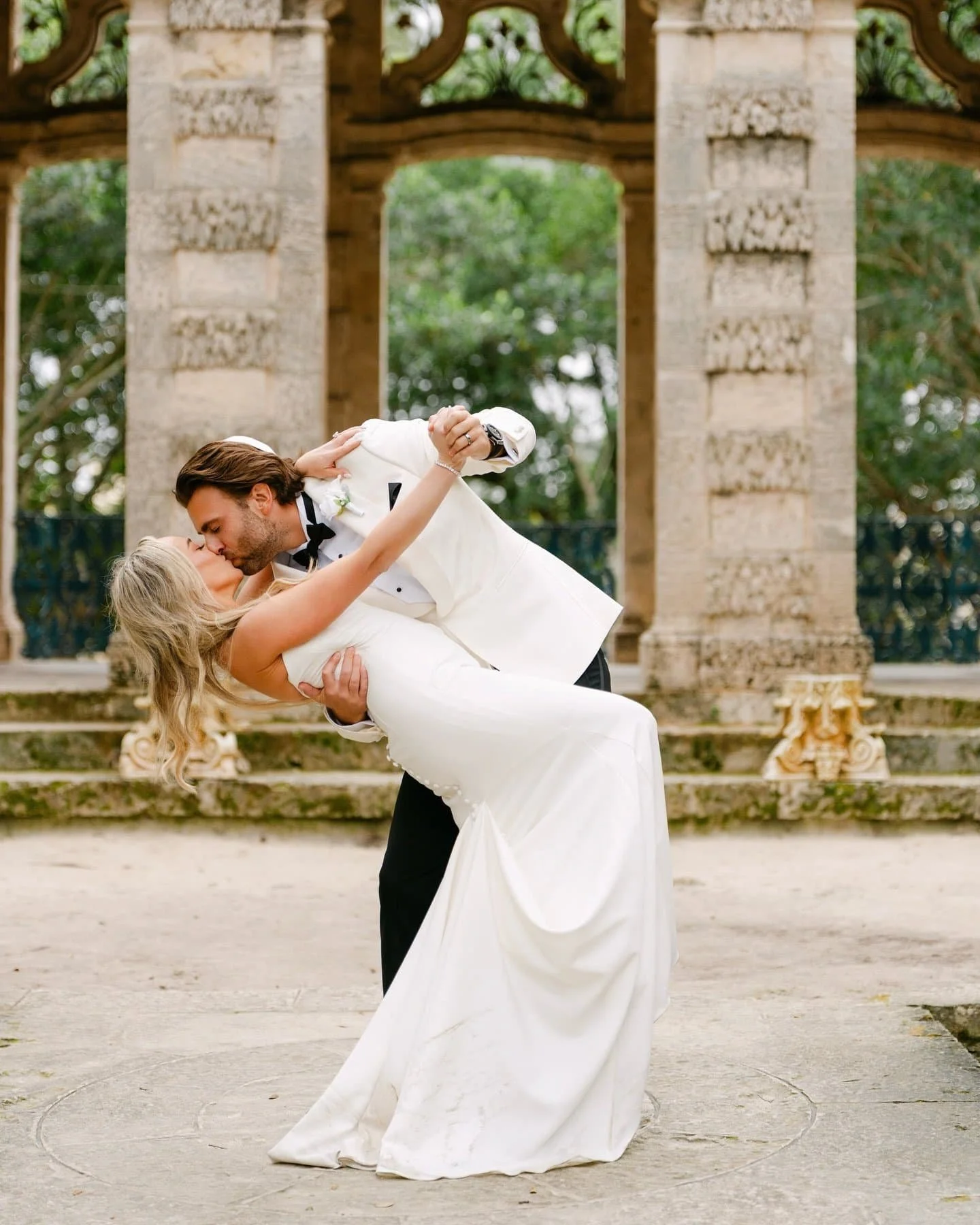 A man in a tuxedo and woman in a wedding dress sharing a kiss while dancing outdoors in front of classical stone architecture.