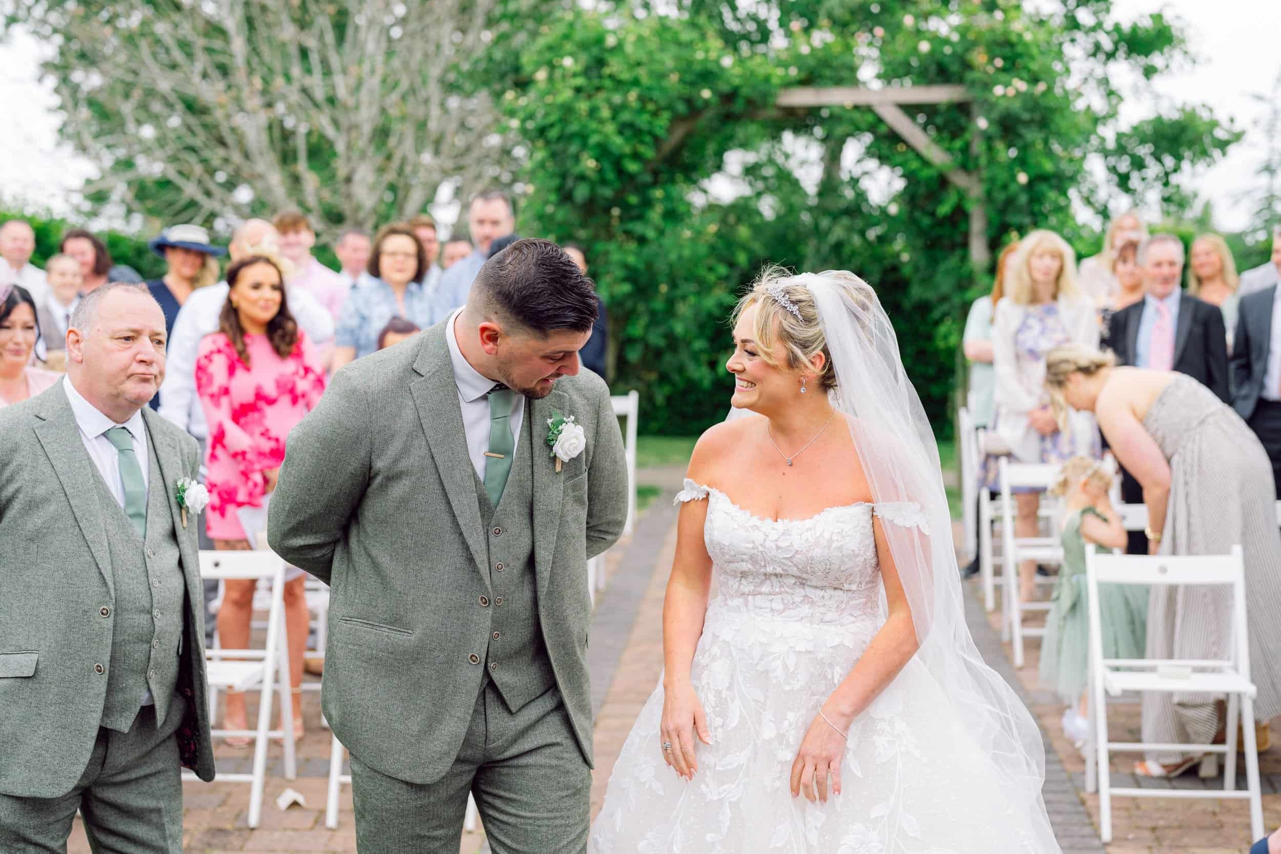 A bride and groom during their outdoor wedding ceremony, smiling at each other, surrounded by seated guests and greenery.