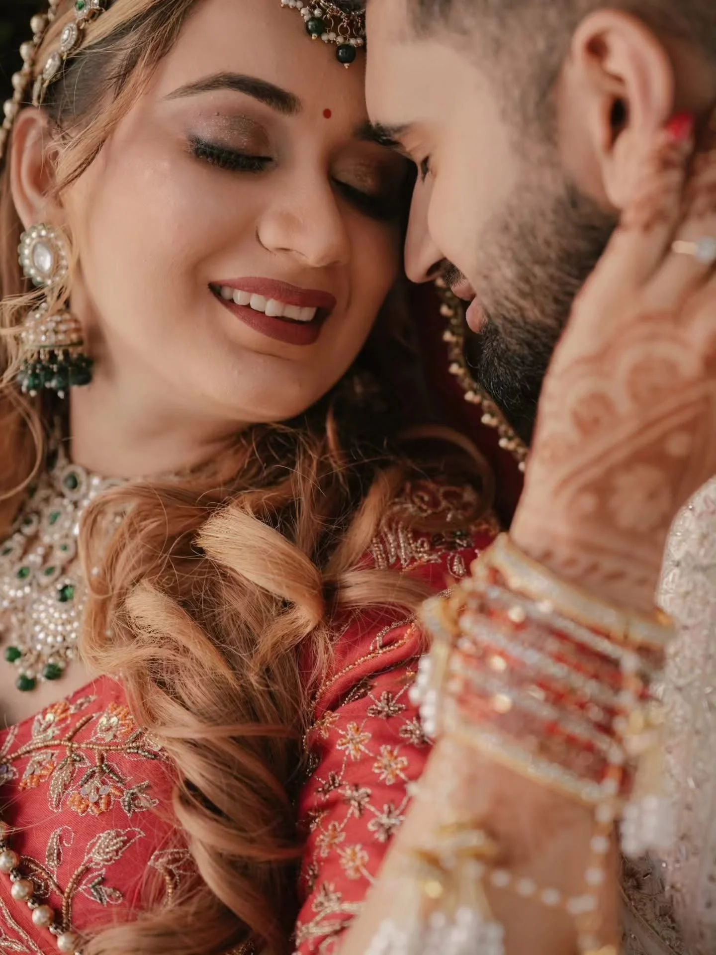 Close-up of a bride and groom with their foreheads touching, smiling with eyes closed, dressed in traditional Indian wedding attire, adorned with jewelry and intricate embroidery.