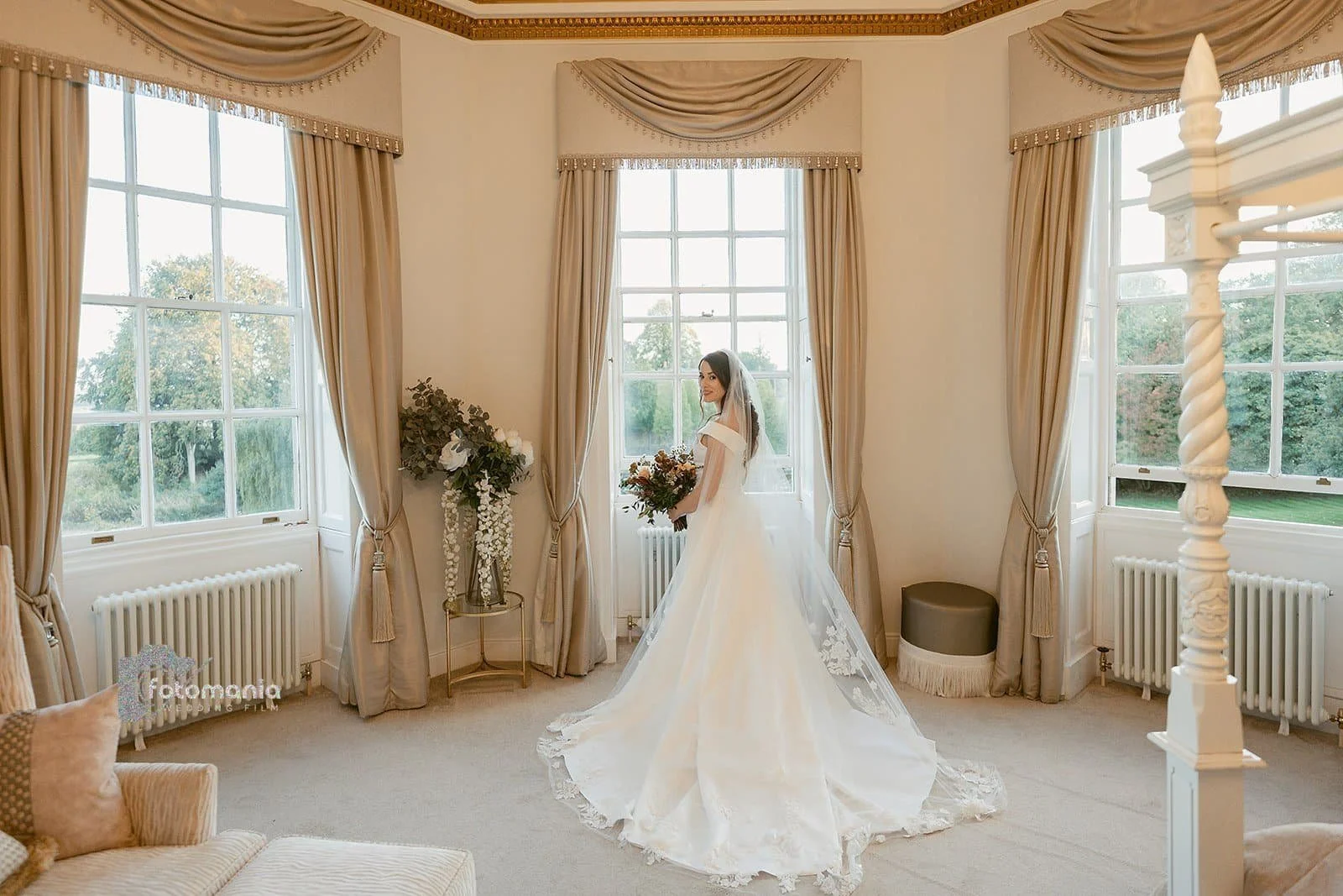 Bride in wedding gown standing in a bright room with large windows, beige curtains, a flower arrangement, and a vintage-style interior.