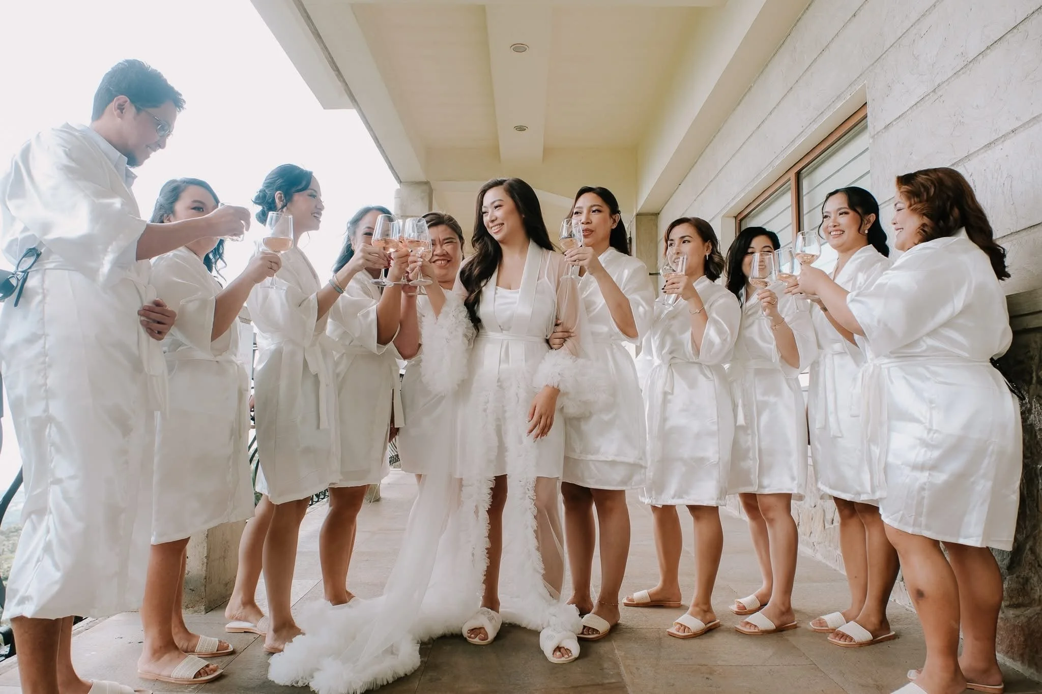 Bride and seven bridesmaids all dressed in matching white robes, holding wine glasses, standing together outside near a building, celebrating a wedding.