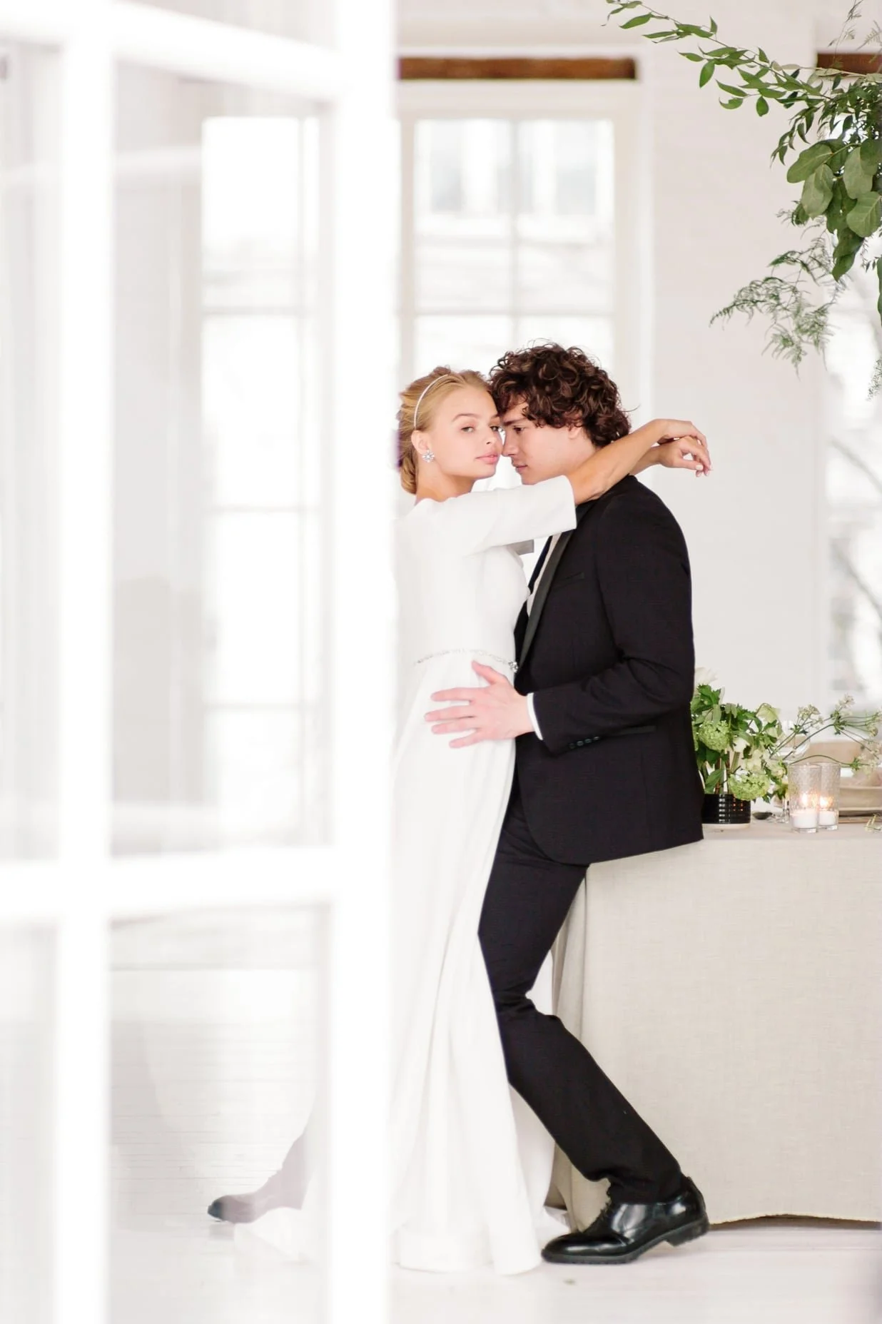 A man and woman in formal attire embracing in a bright, modern room with large windows and white walls, with greenery and candles in the background.