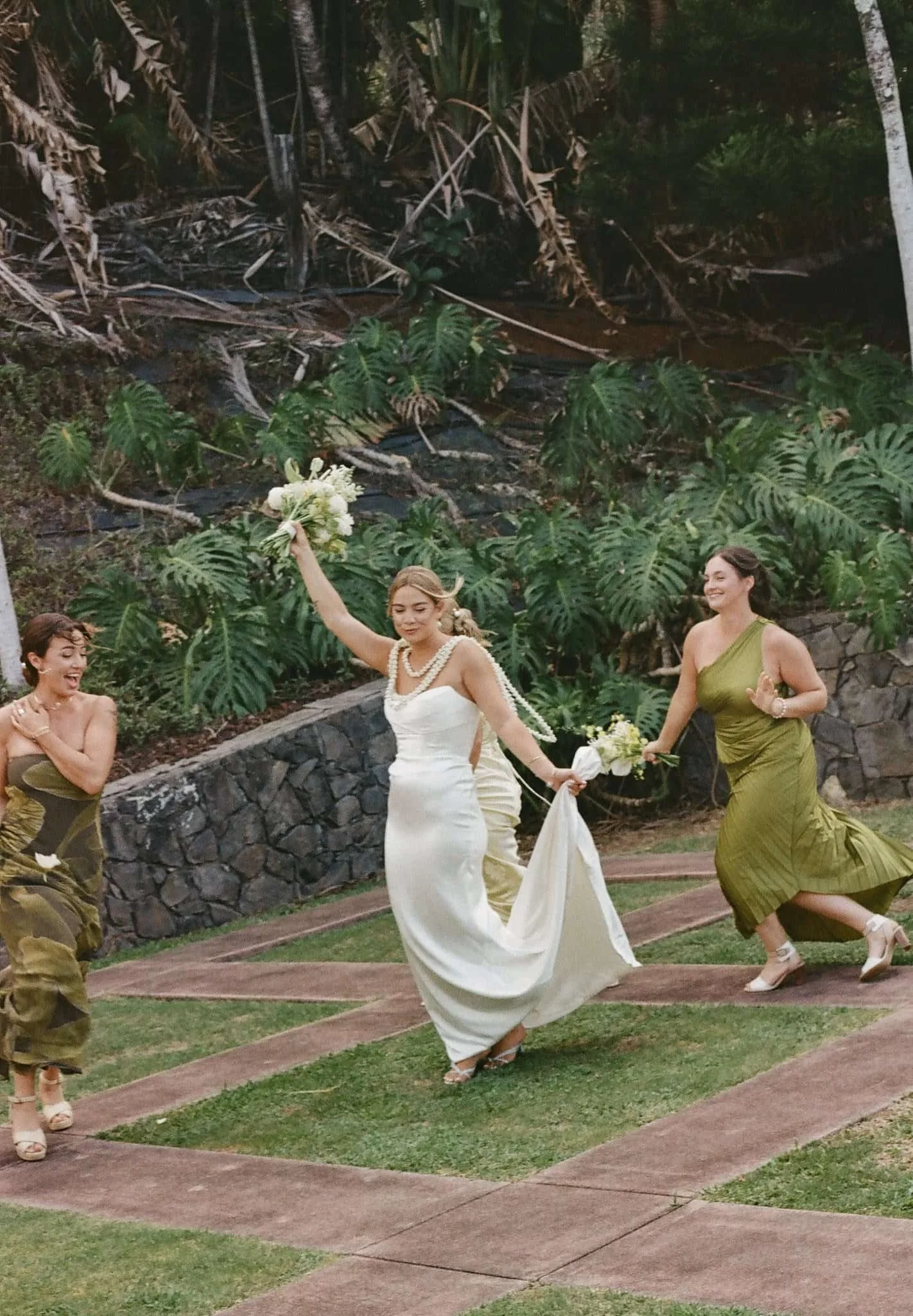 A bride in a white dress holding a bouquet, dancing in a garden, surrounded by women in green dresses, during a wedding celebration.