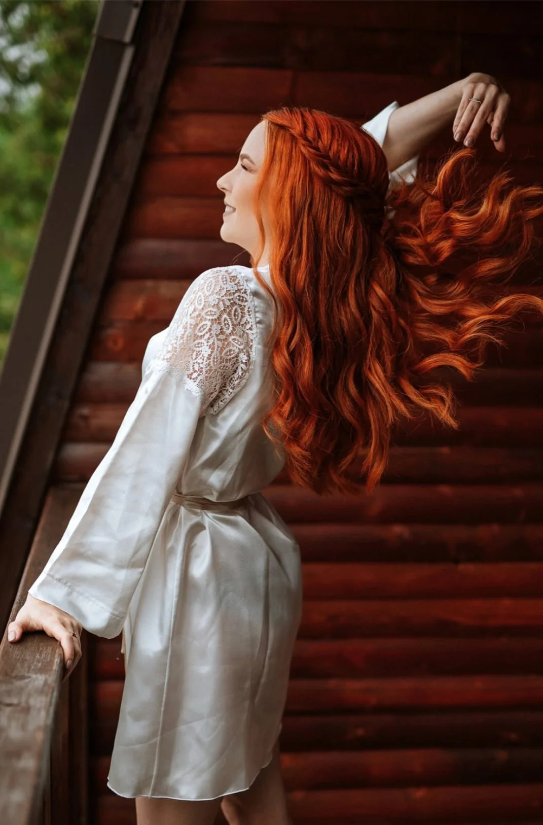 Woman with long, wavy red hair leaning against a wooden railing, smiling with eyes closed, wearing a white silk robe with lace details on the sleeves, standing outdoors near a wooden wall.