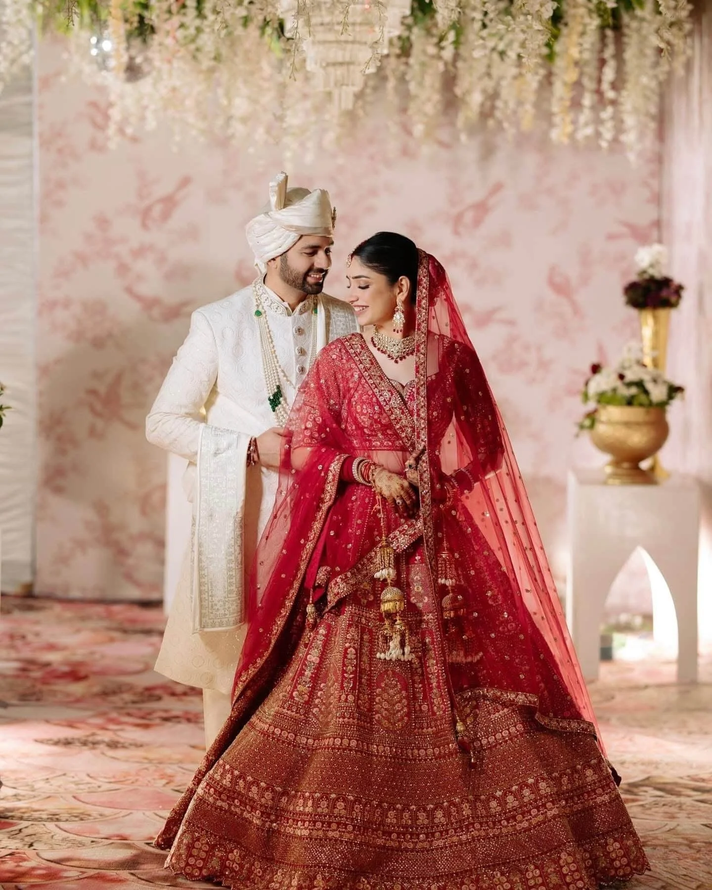 A bride and groom dressed in traditional Indian wedding attire, standing close together and smiling. The bride is wearing a red richly embroidered lehenga with jewelry and a red veil, while the groom is wearing a cream-colored sherwani with a turban 