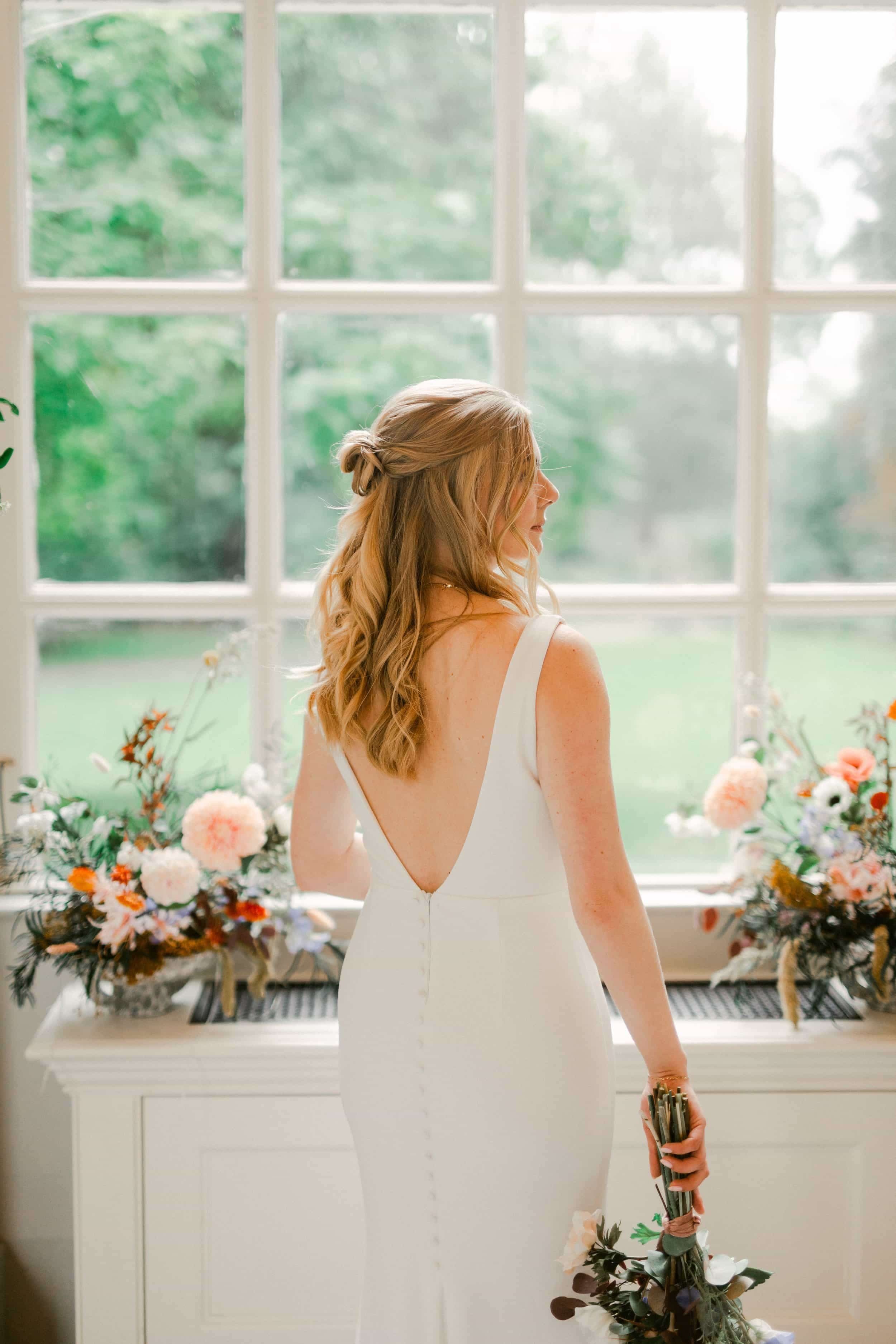 A woman with long blond hair styled in loose waves is standing near a window with a garden view. She is wearing a white dress with a low back, holding a bouquet of flowers with various blooms and greenery.