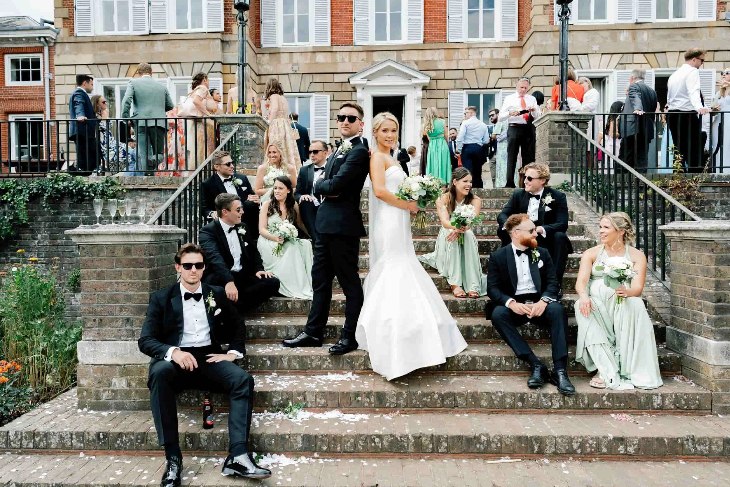 A wedding celebration on outdoor steps in front of a brick building with white window shutters. The bride and groom are standing in the center, surrounded by wedding guests dressed in formal attire, some seated and others standing.